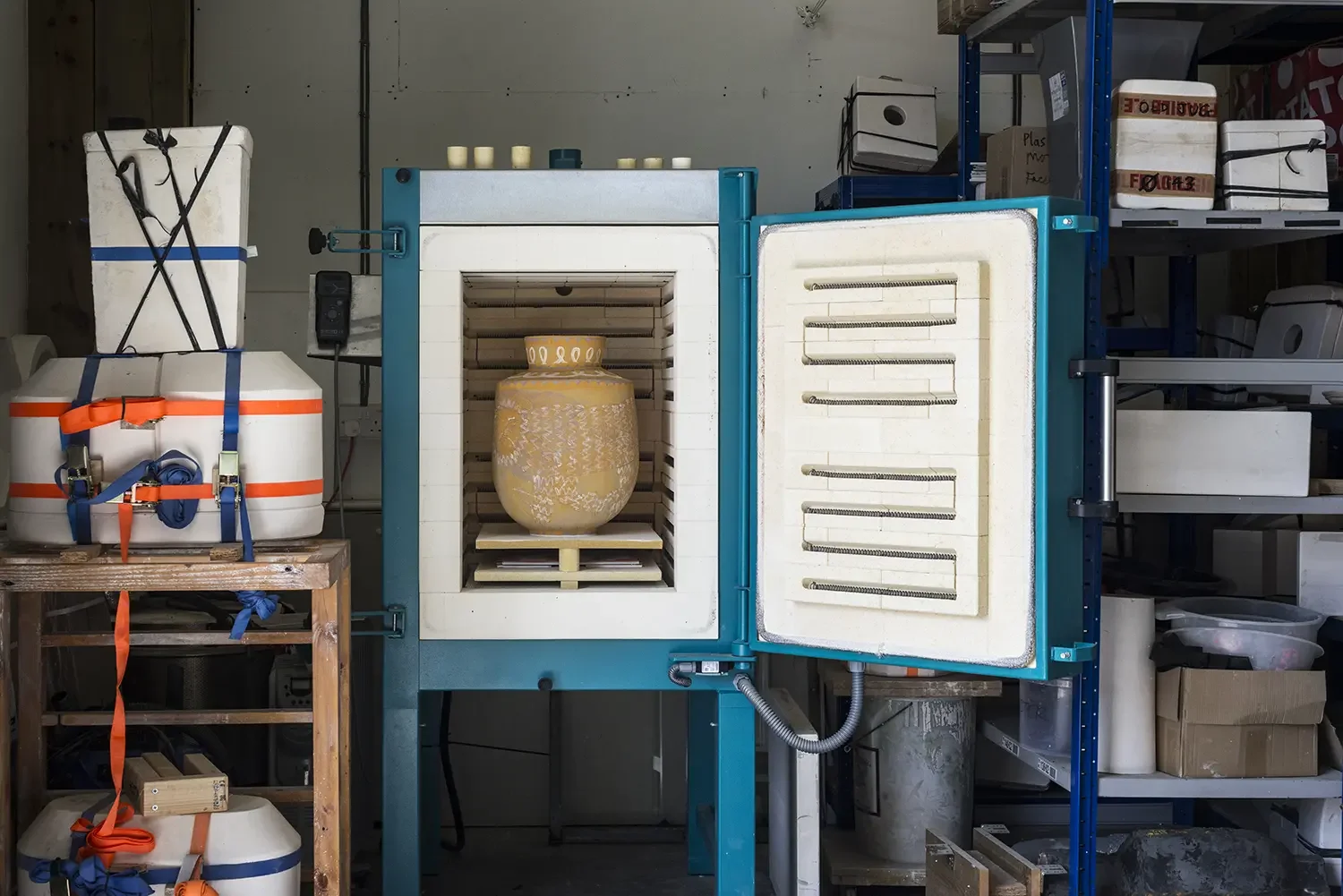 A kiln with a decorative vase inside, located in an industrial or art studio, surrounded by shelves and storage containers.