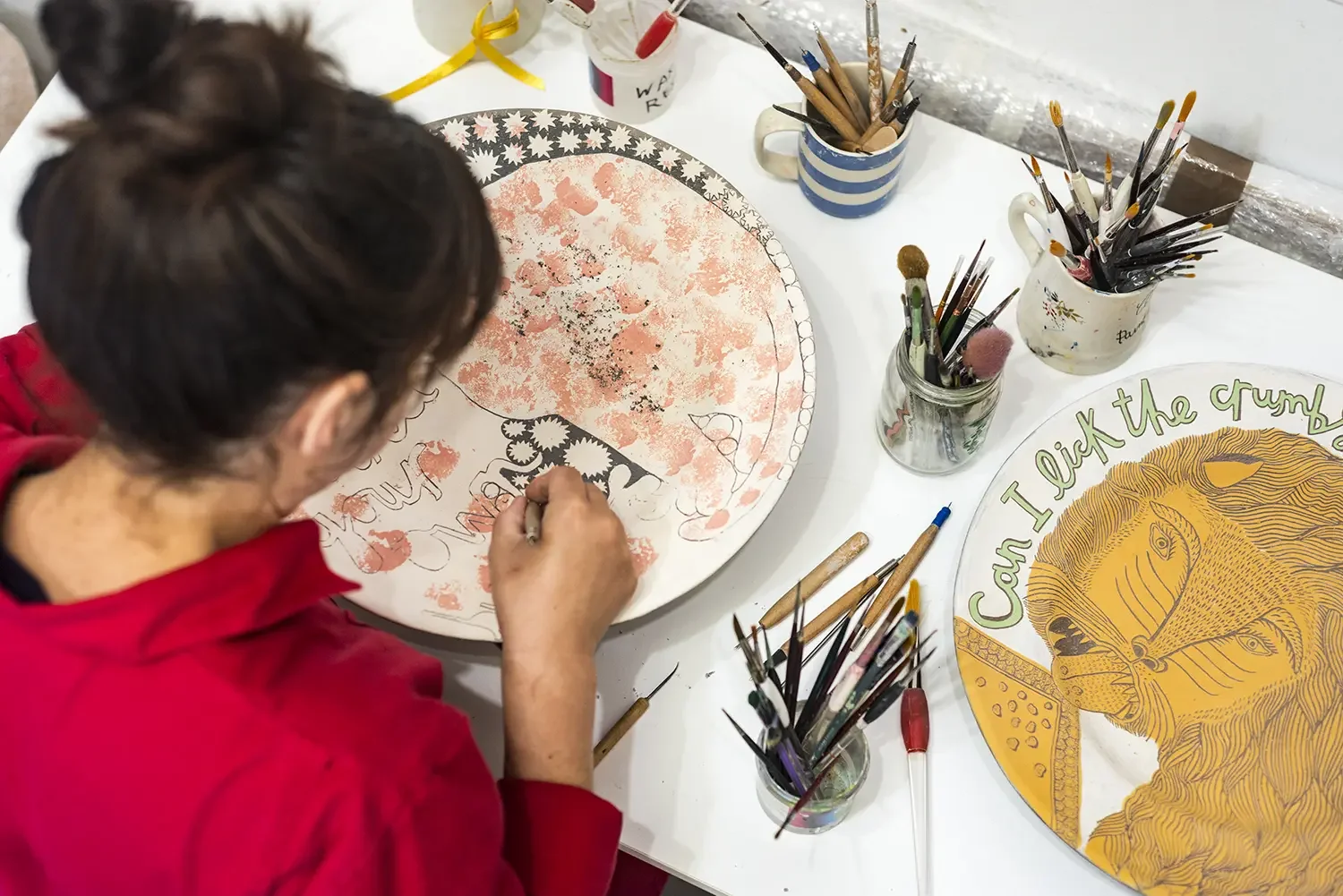 Vicky Lindo working on a circular plate with orange and black details, surrounded by tools used for her sgraffito on a white table.