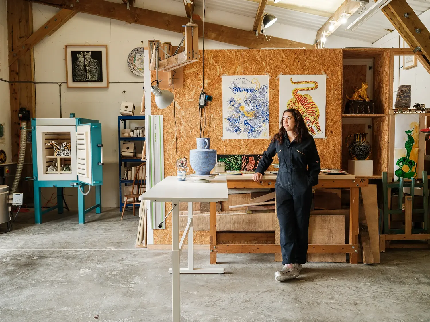 Vicky Lindo in black overalls leaning on a wooden worktable inside her art studio with paintings and sculptures displayed on the walls and shelves.