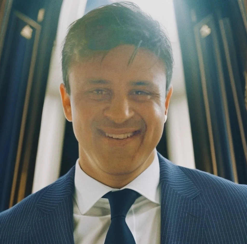 A young man in a navy pinstripe suit, white shirt, and dark tie, smiling confidently, with dark hair and a bright background.