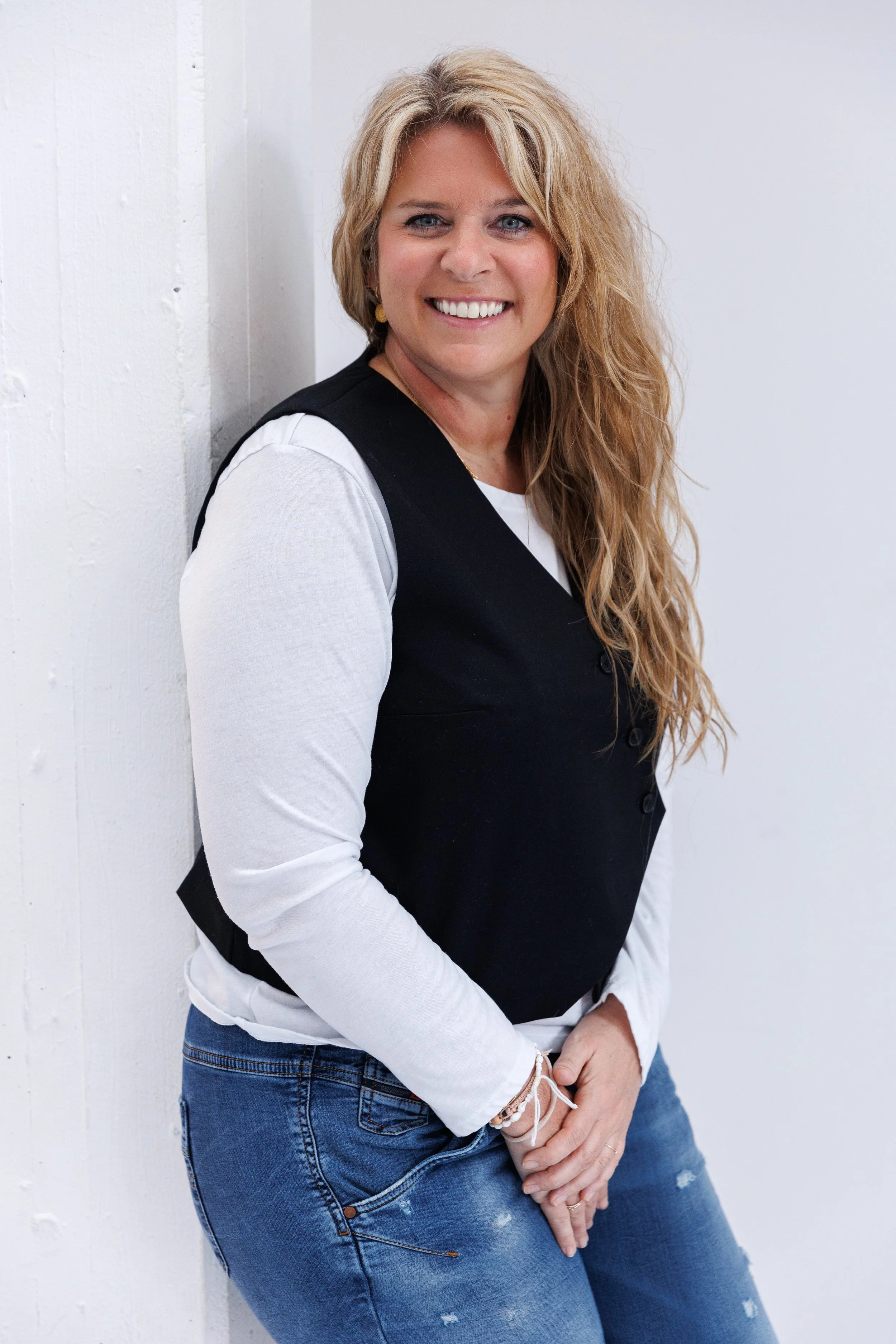 Eine lächelnde Frau mit langen, gewellten blonden Haaren trägt eine schwarze Weste über einem weißen Langarmshirt und blaue Jeans, steht an einer weißen Wand und lächelt in die Kamera.