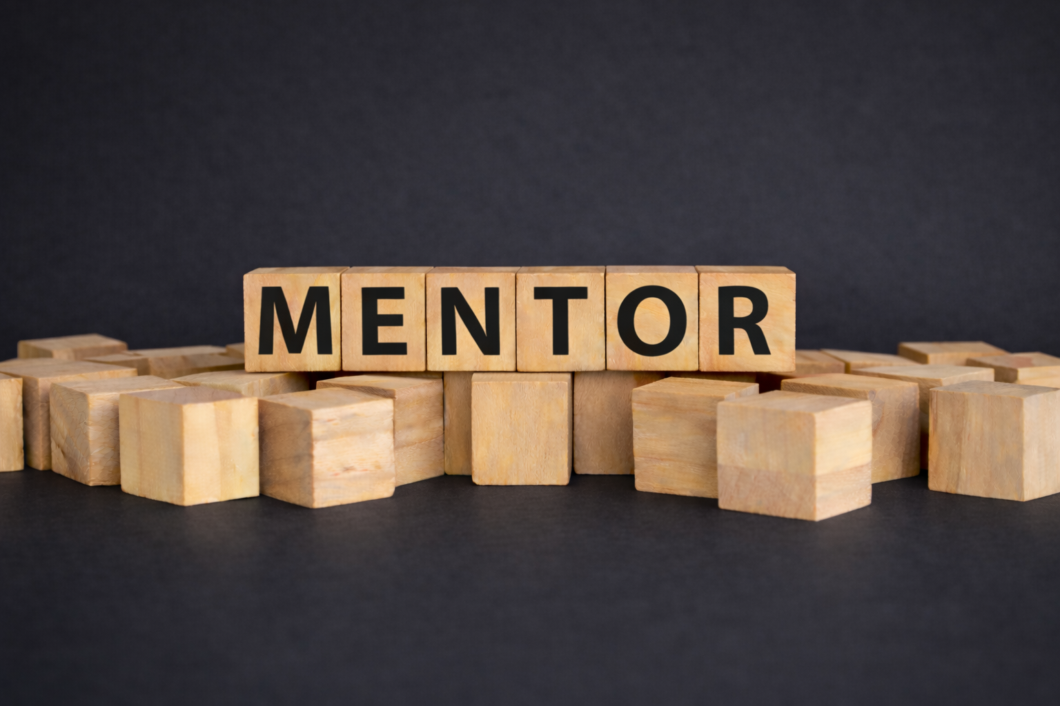 Holzbuchstaben, die das Wort 'MENTOR' bilden, auf einer schwarzen Oberfläche mit zusätzlichen Holzblöcken im Hintergrund.
