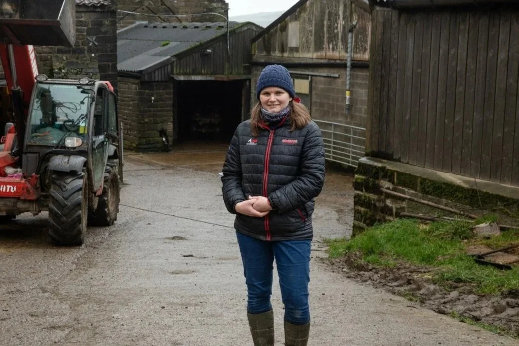 A young woman standing outdoors in a rural or farm setting, wearing a dark jacket, blue jeans, a knit hat, and rain boots, smiling at the camera.
