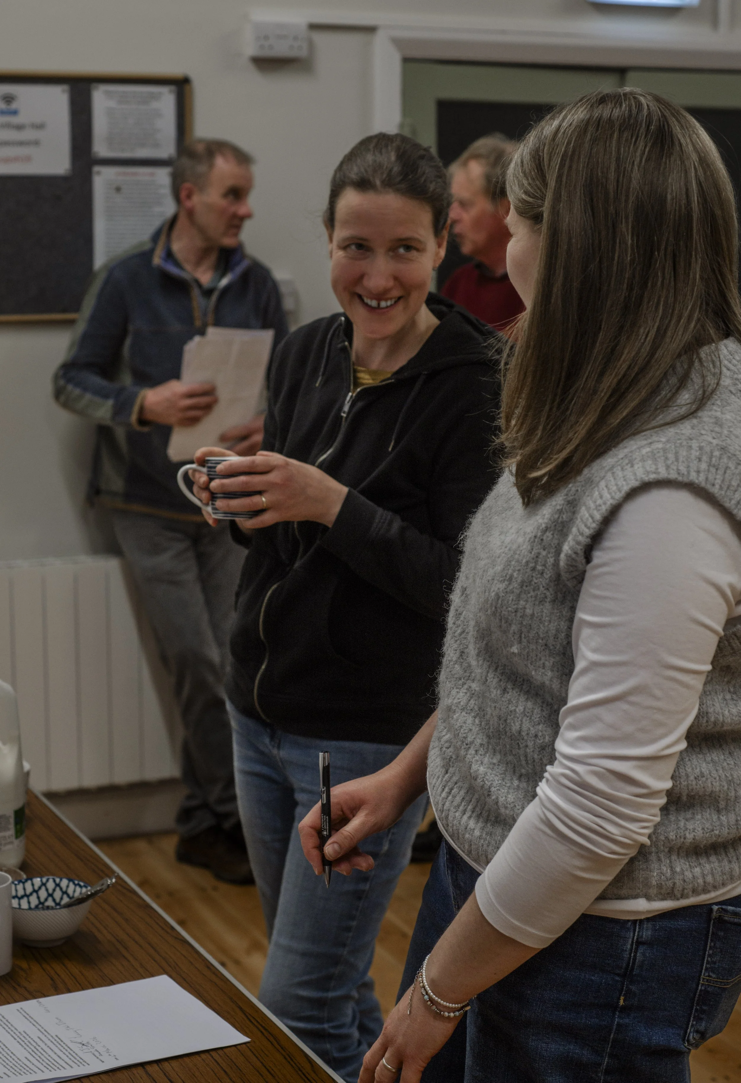 Four people are gathered at a table, two women in focus engaged in conversation, while two men in the background hold papers, in a room with a white wall and bulletin board.