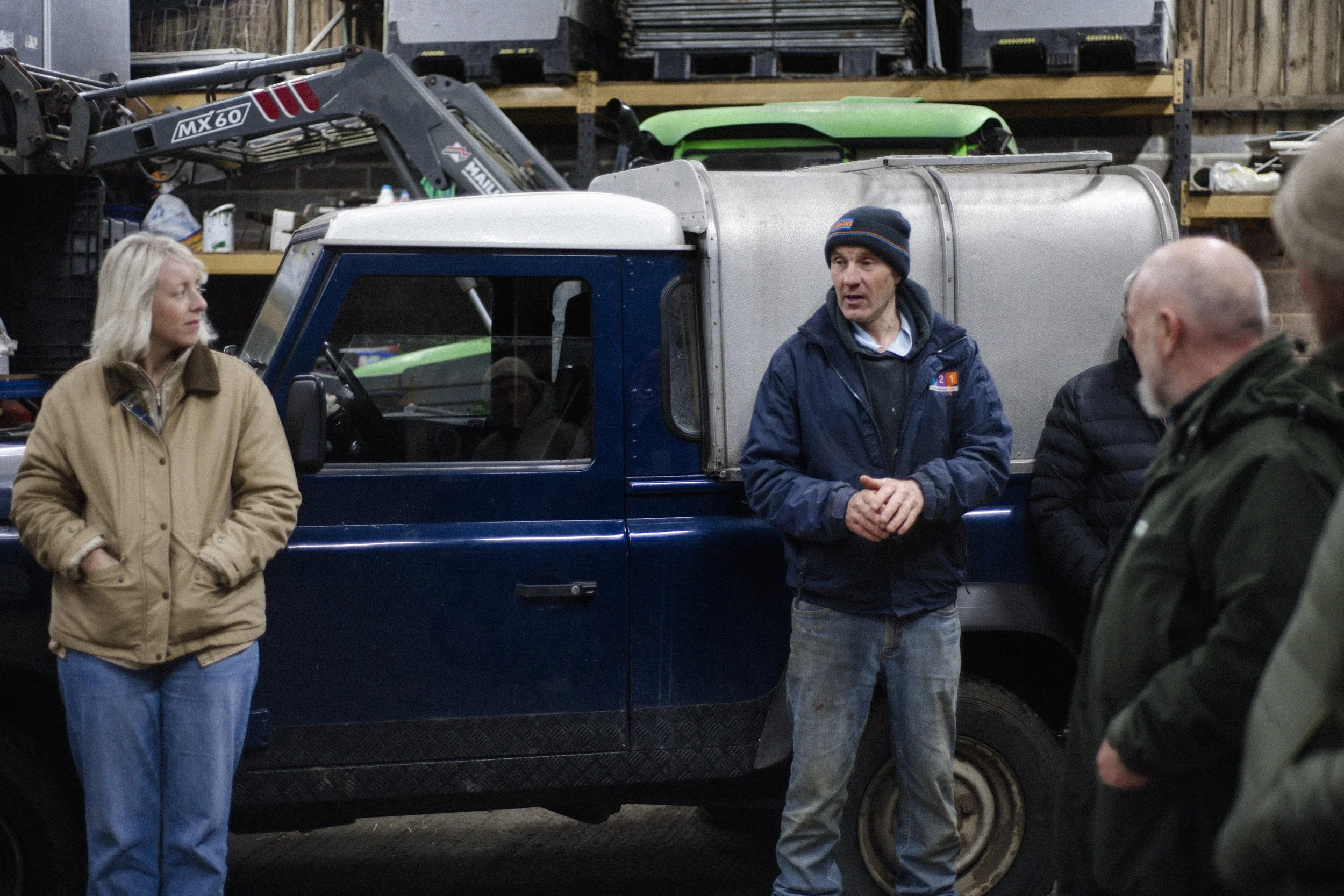 Group of people standing near a blue work truck inside a workshop or garage, with one man speaking to others, surrounded by shelves with tools and equipment.