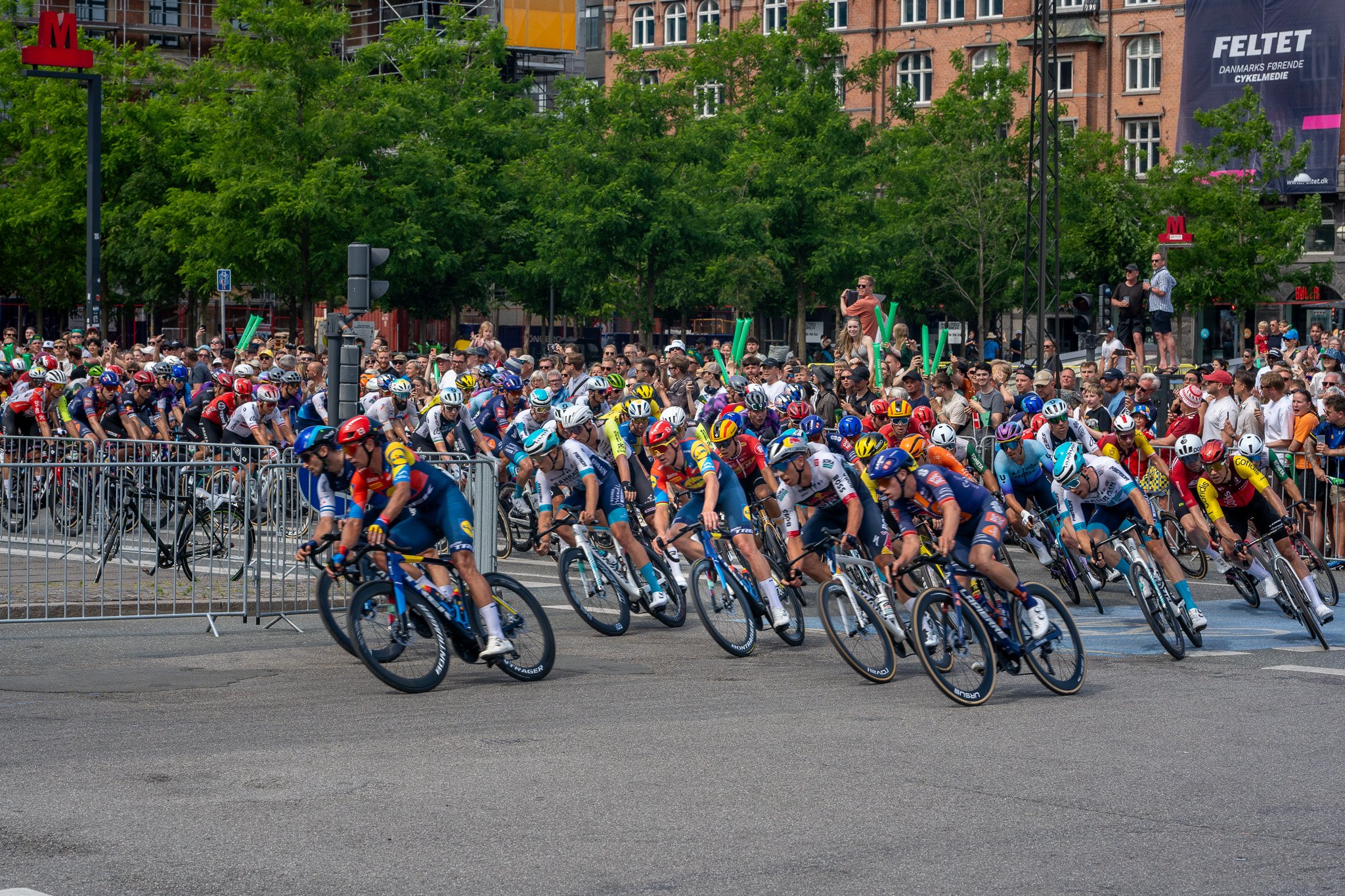 Cyclists participating in a race, leaning into a turn on city streets, with spectators watching behind metal barriers and some cheering.