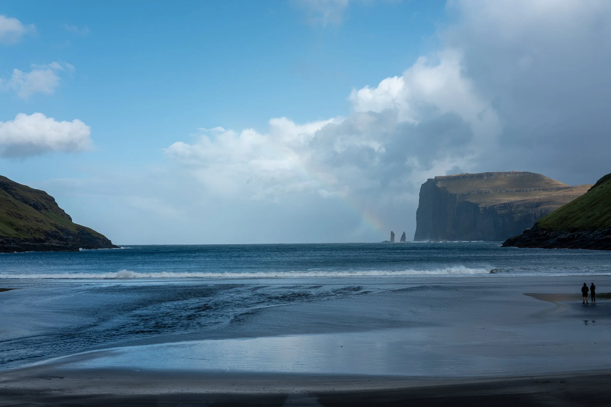 A scenic coastal landscape featuring a calm ocean, rocky cliffs on both sides, a cloudy sky with patches of blue, a faint rainbow near the horizon, and two people walking along the beach.