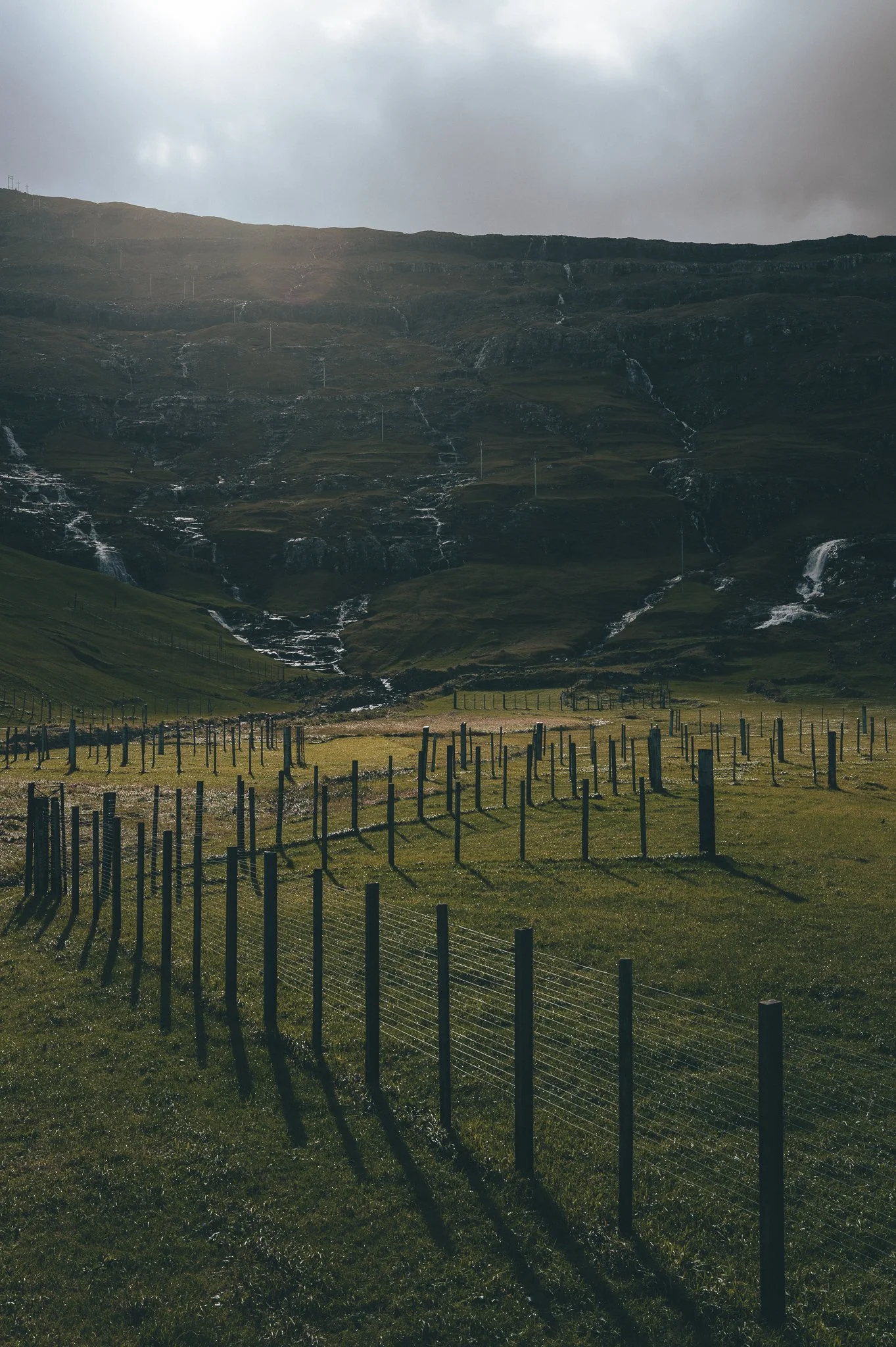A grassy field in the foreground with a wire fence. In the background, a mountain with multiple small streams or waterfalls flowing down its slopes, under a cloudy sky.