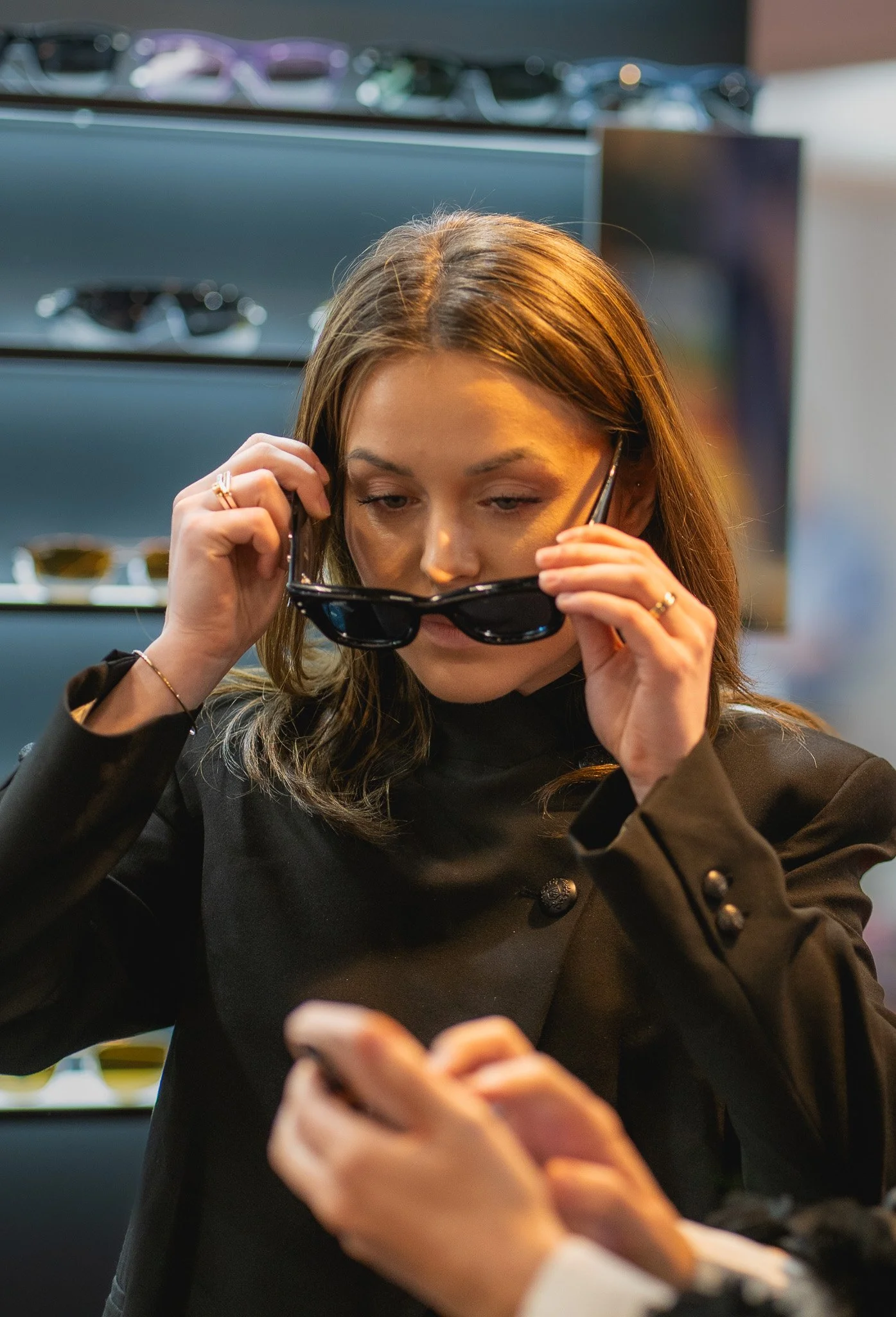A woman with light brown hair, wearing a black blazer, is holding sunglasses in one hand and looking at her smartphone in an indoor setting, with a display of sunglasses in the background.