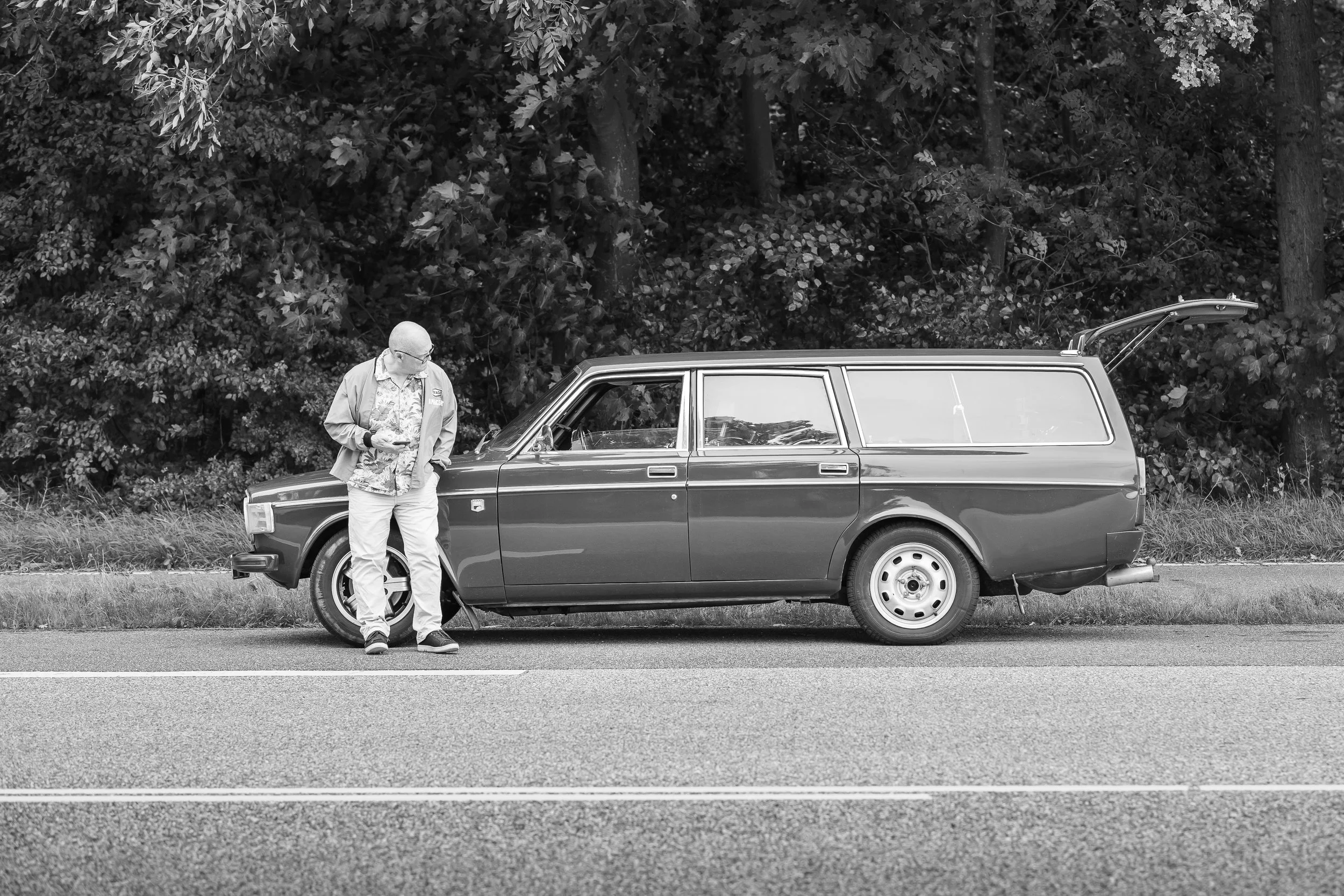 A man standing next to a vintage station wagon on the side of a two-lane road, with a forested background.