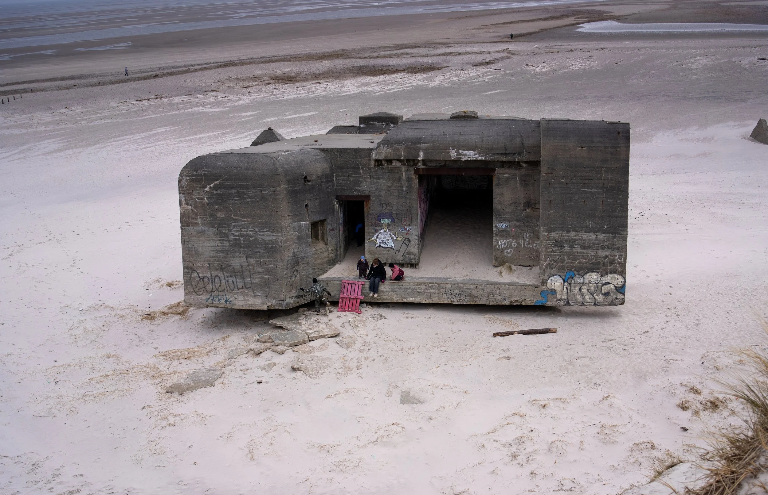 Concrete bunker on sandy beach with graffiti, small group of children sitting outside, wooden pallet and fallen wood nearby, overcast sky.