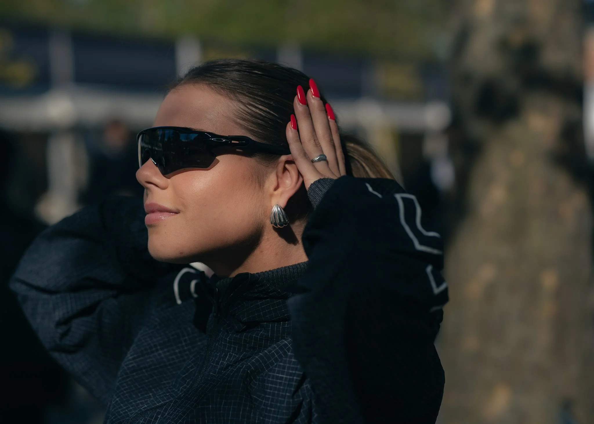 Woman with dark hair in a ponytail, wearing black sunglasses, silver earrings, and a dark jacket with white stripes, adjusting her hair outdoors with blurred background.