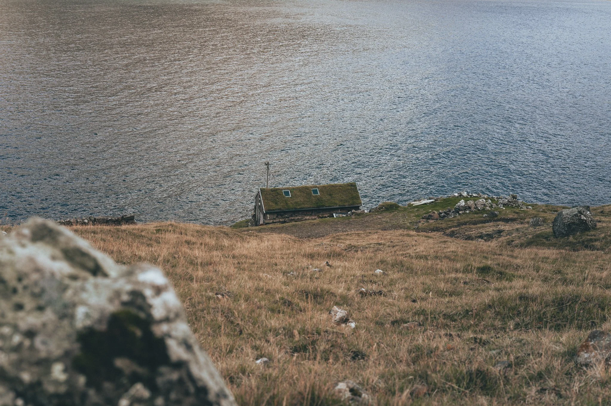 A house with a moss-covered roof situated on a grassy hill near a body of water.