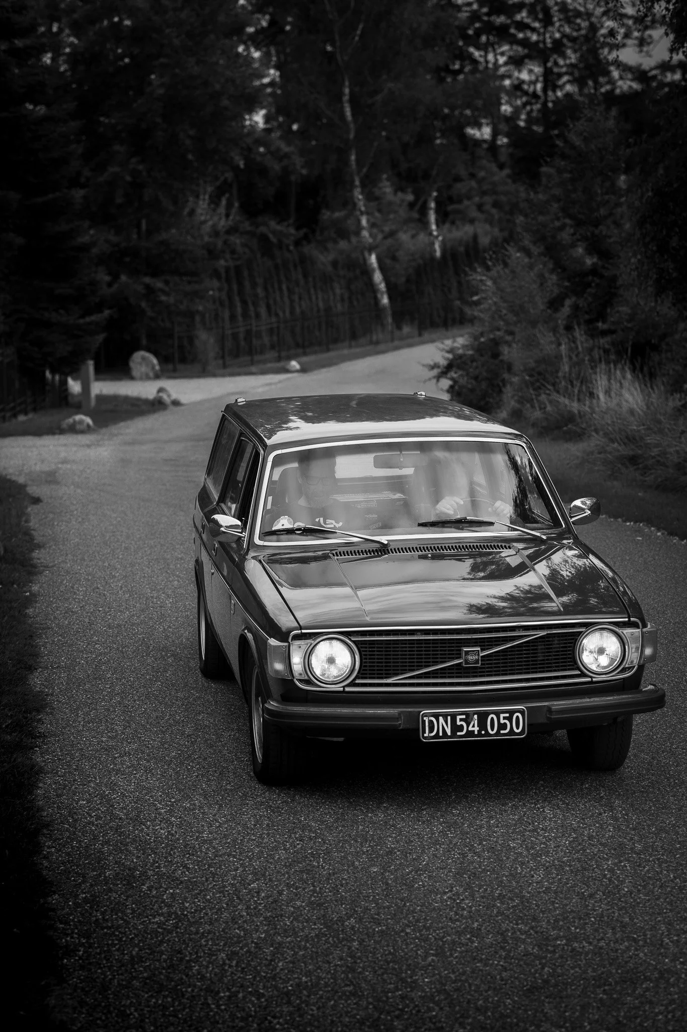 A black and white photo of an old car driving on a winding road surrounded by trees and rocks.