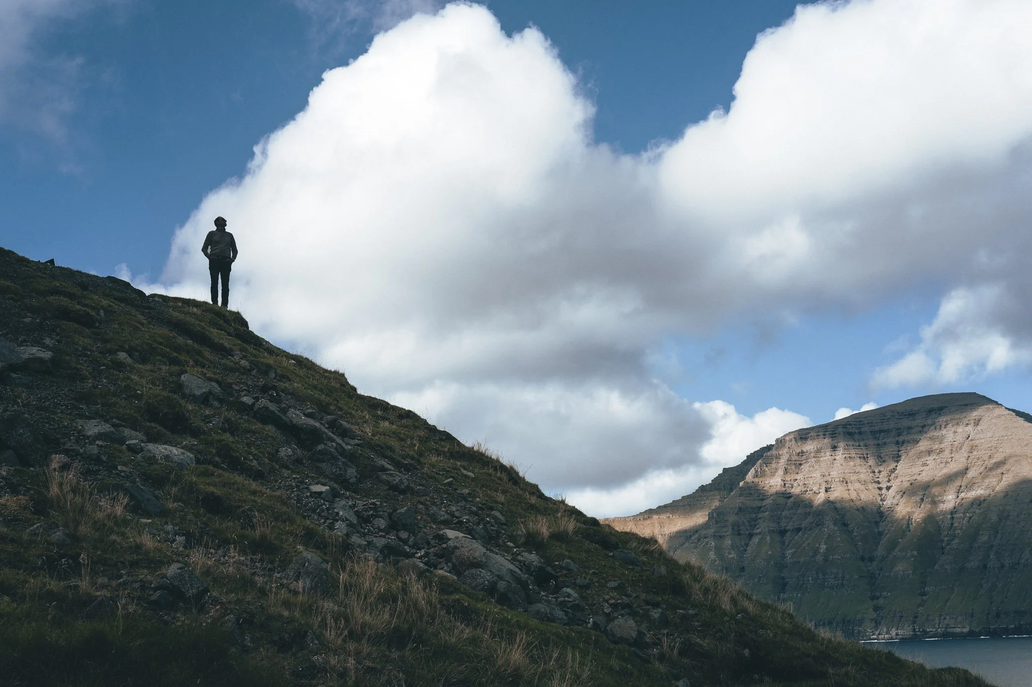 A person standing on a grassy, rocky hillside overlooking a mountain landscape with clouds and a partly cloudy sky.