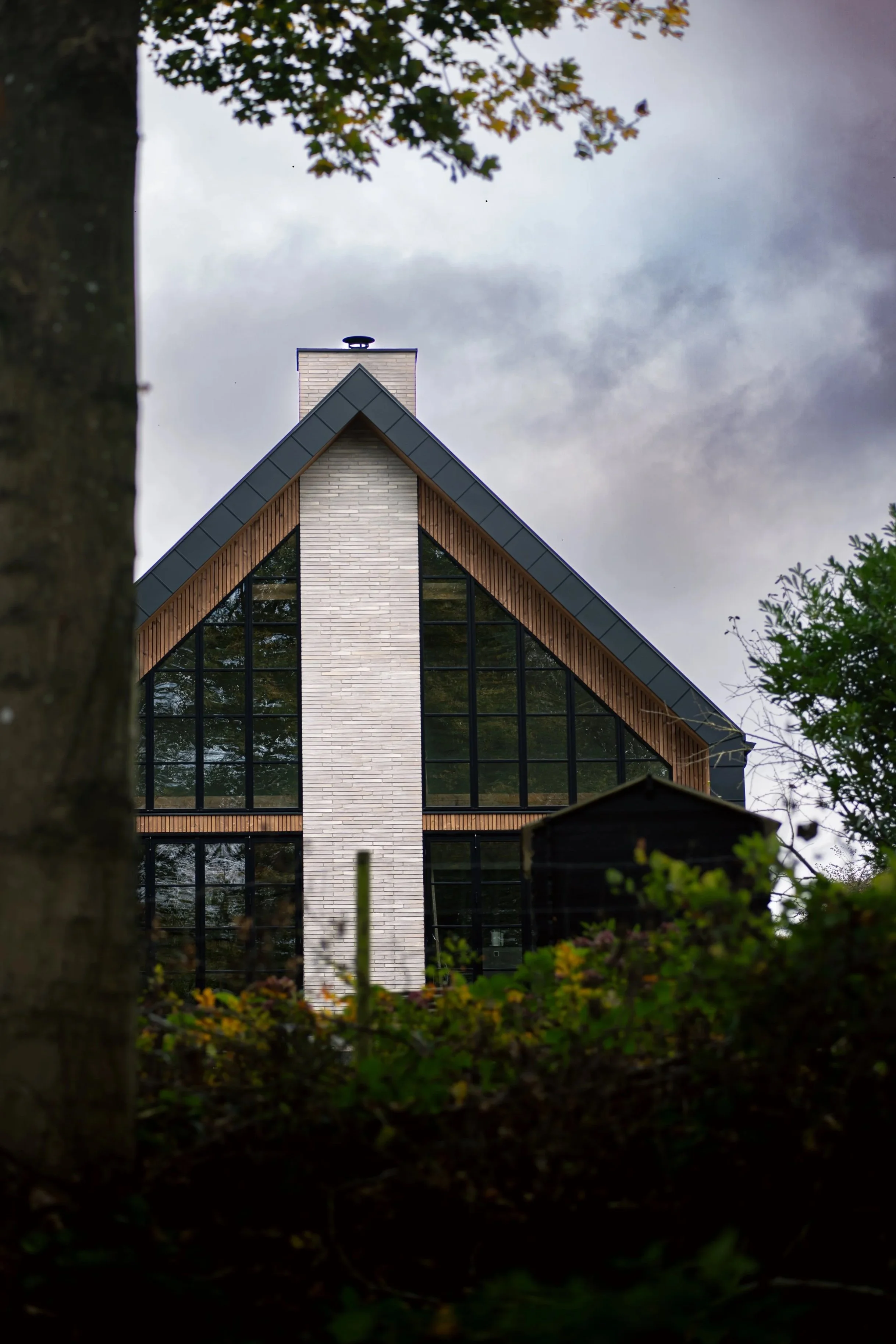 Modern Scandinavian architecture featuring a minimalist gabled house with large glass windows, surrounded by nature under a dramatic sky.