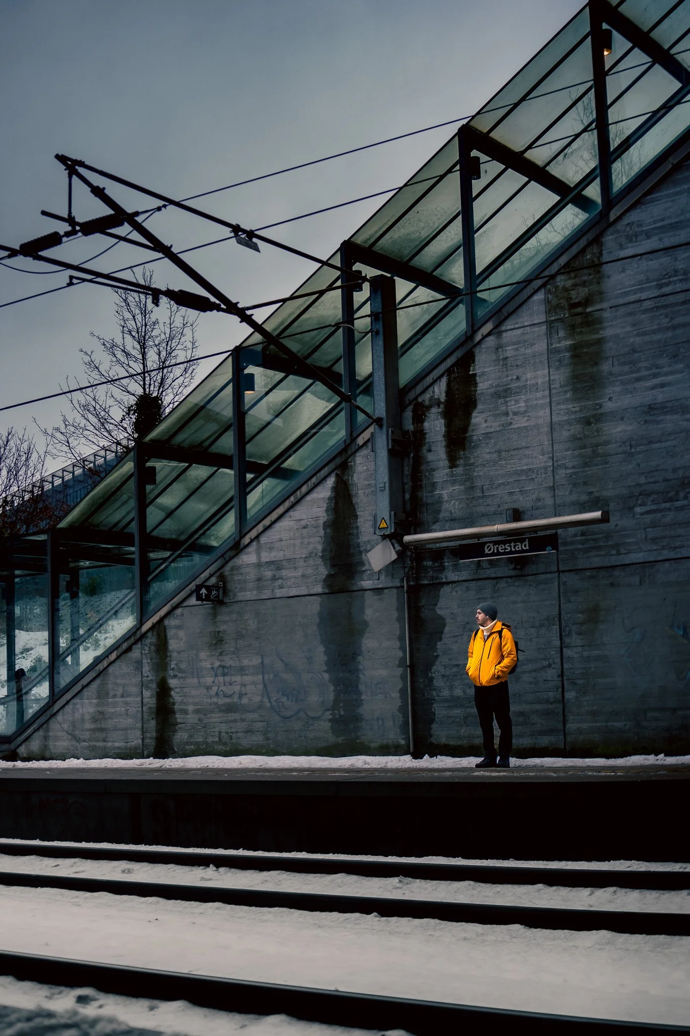 A person in a yellow jacket standing on a train platform at Orestad train station in Denmark, with snow on the ground and an inclined glass-walled staircase leading up to an overhead walkway.