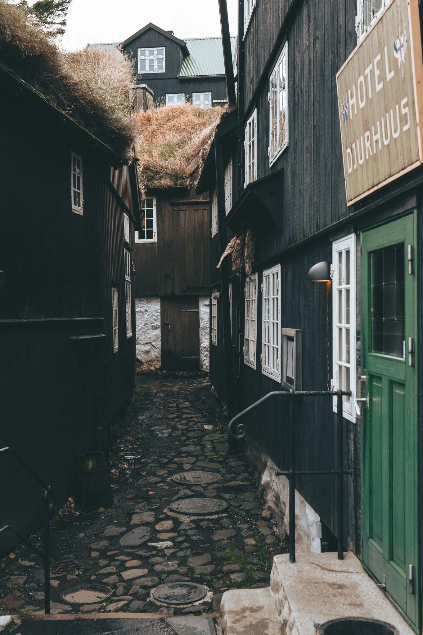 A narrow cobblestone alley between black wooden buildings with white-framed windows. A green door is visible on the right, and a sign with 'HOTEL DJURHUIS' is on the upper right. There are some plants and a hillside with buildings at the top of the a