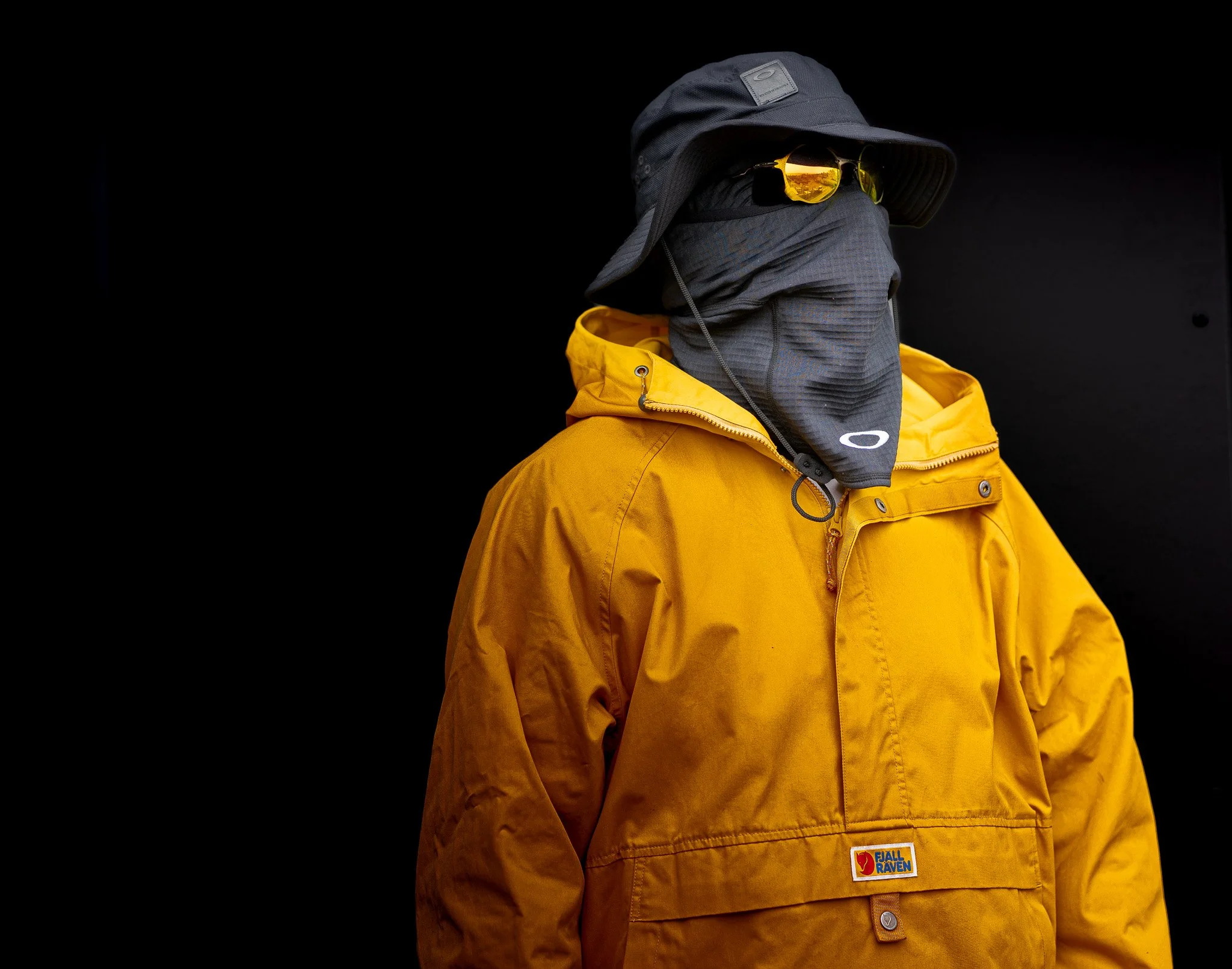 A mannequin dressed in yellow outdoor jacket, black hat, sunglasses, and face covering against a black background.
