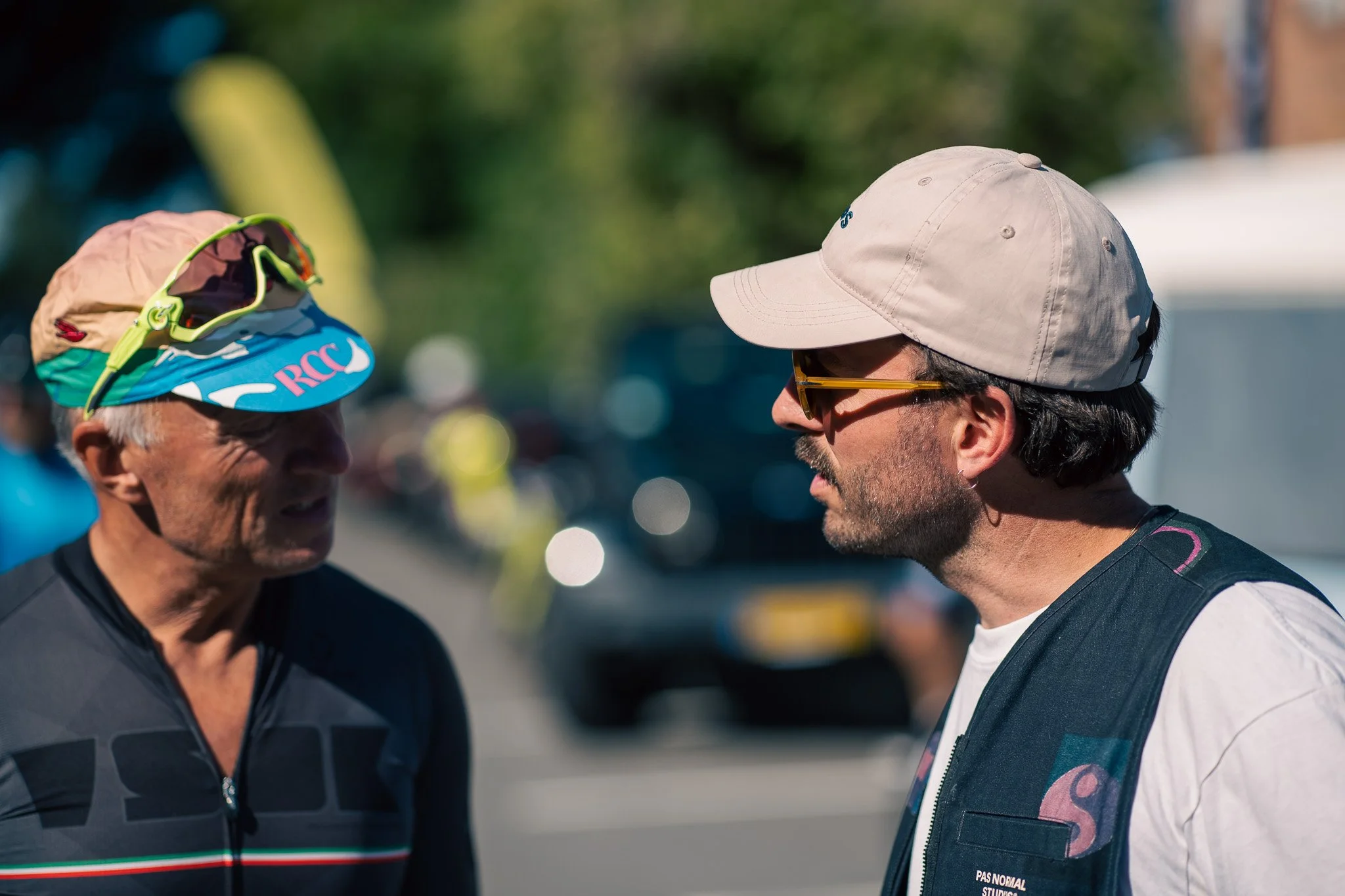 Two men are having a conversation outdoors, both facing each other. The man on the left wears a black cycling jersey, a multicolored hat with sunglasses resting on top, and has grey hair. The man on the right wears a light beige baseball cap, sunglas