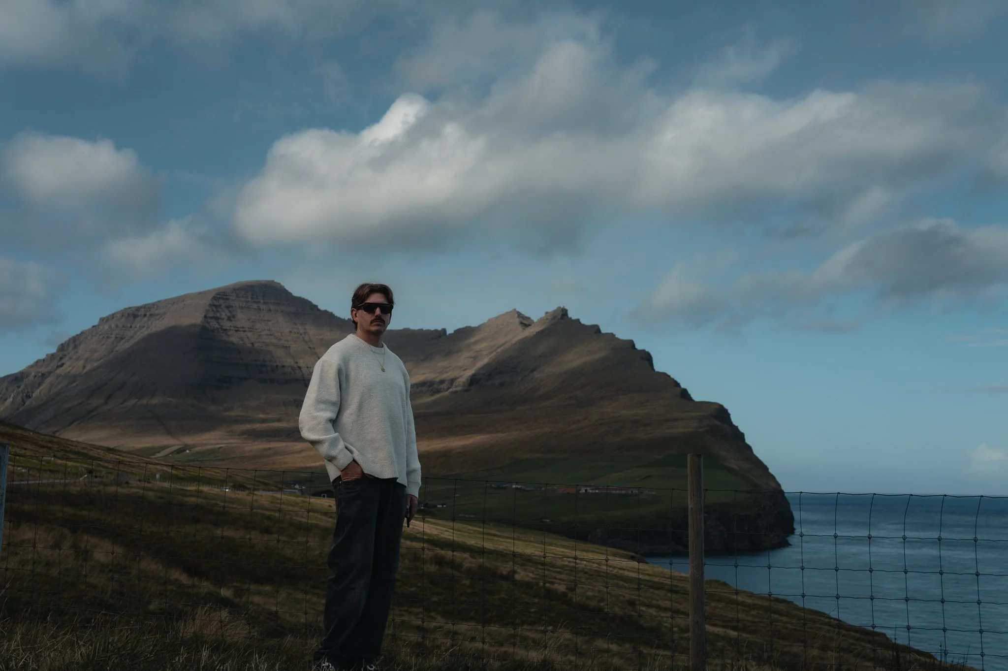 A man wearing sunglasses, a light gray sweater, and dark pants standing outdoors near a fence with mountainous landscape and a cloudy sky in the background.