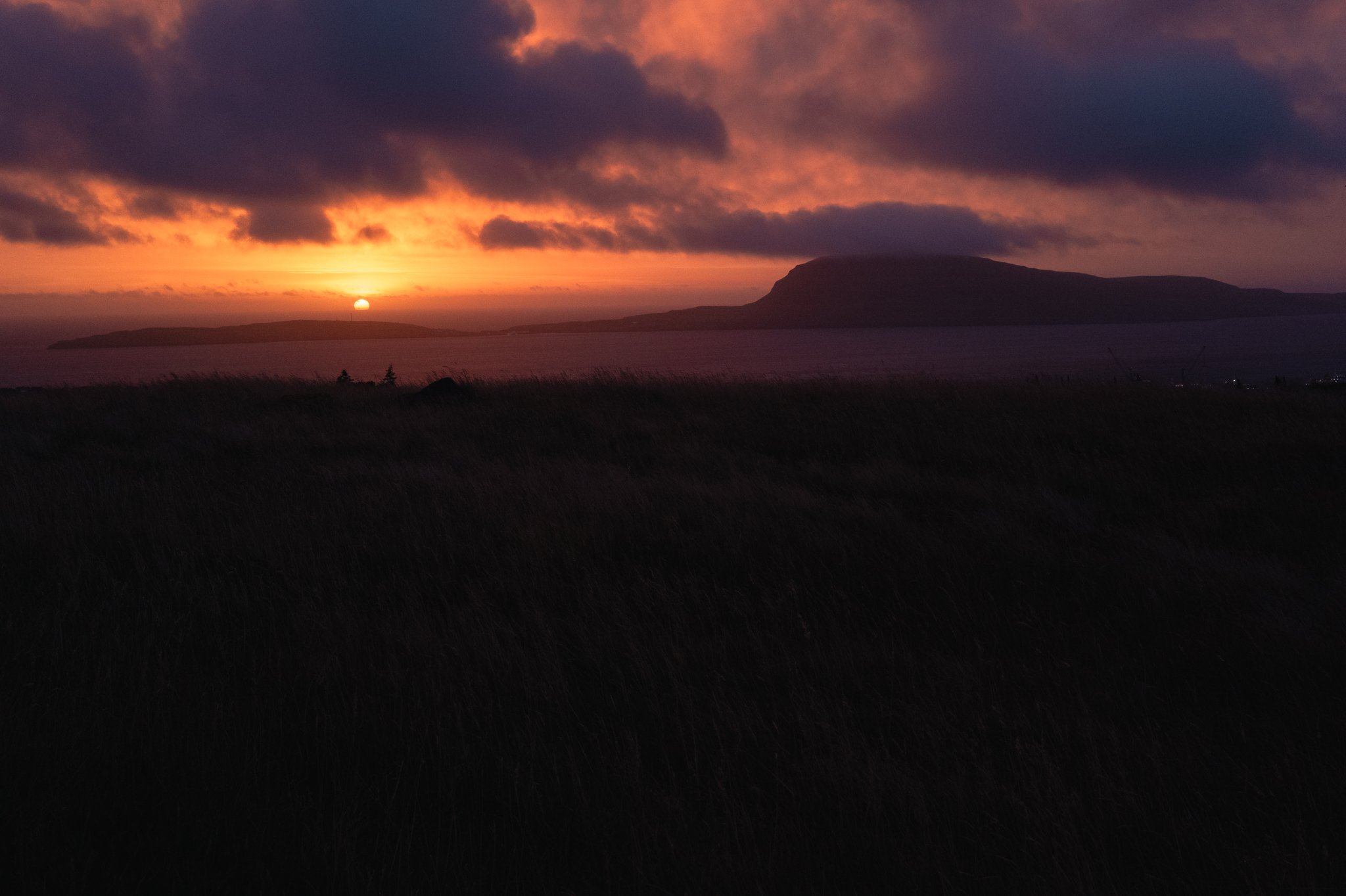 Sun setting over a distant mountain and body of water with dark clouds in the sky, creating a colorful sunset scene.