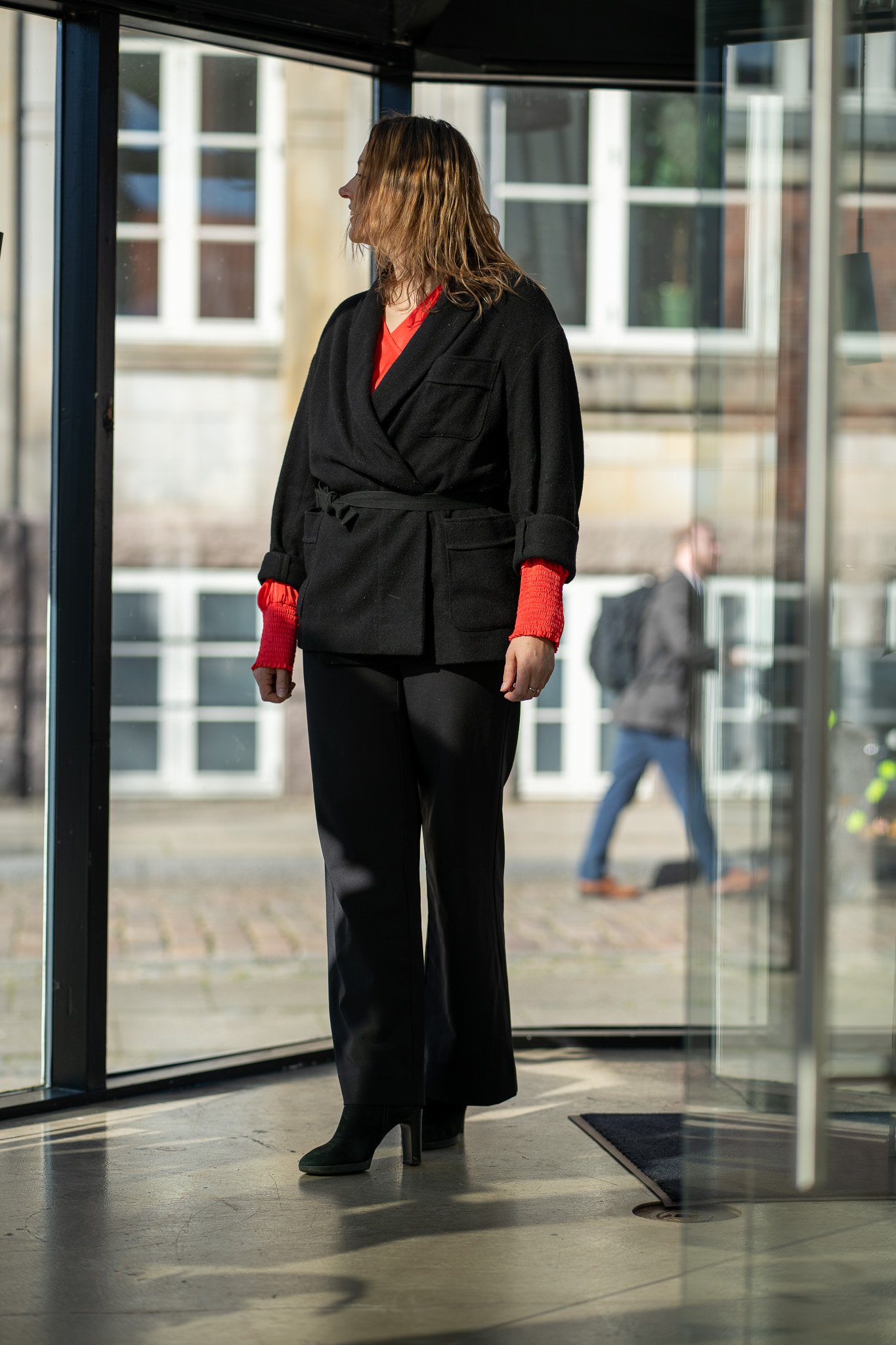A woman with shoulder-length brown hair wearing a black coat, red sweater, black pants, and black heeled boots standing near an entrance.