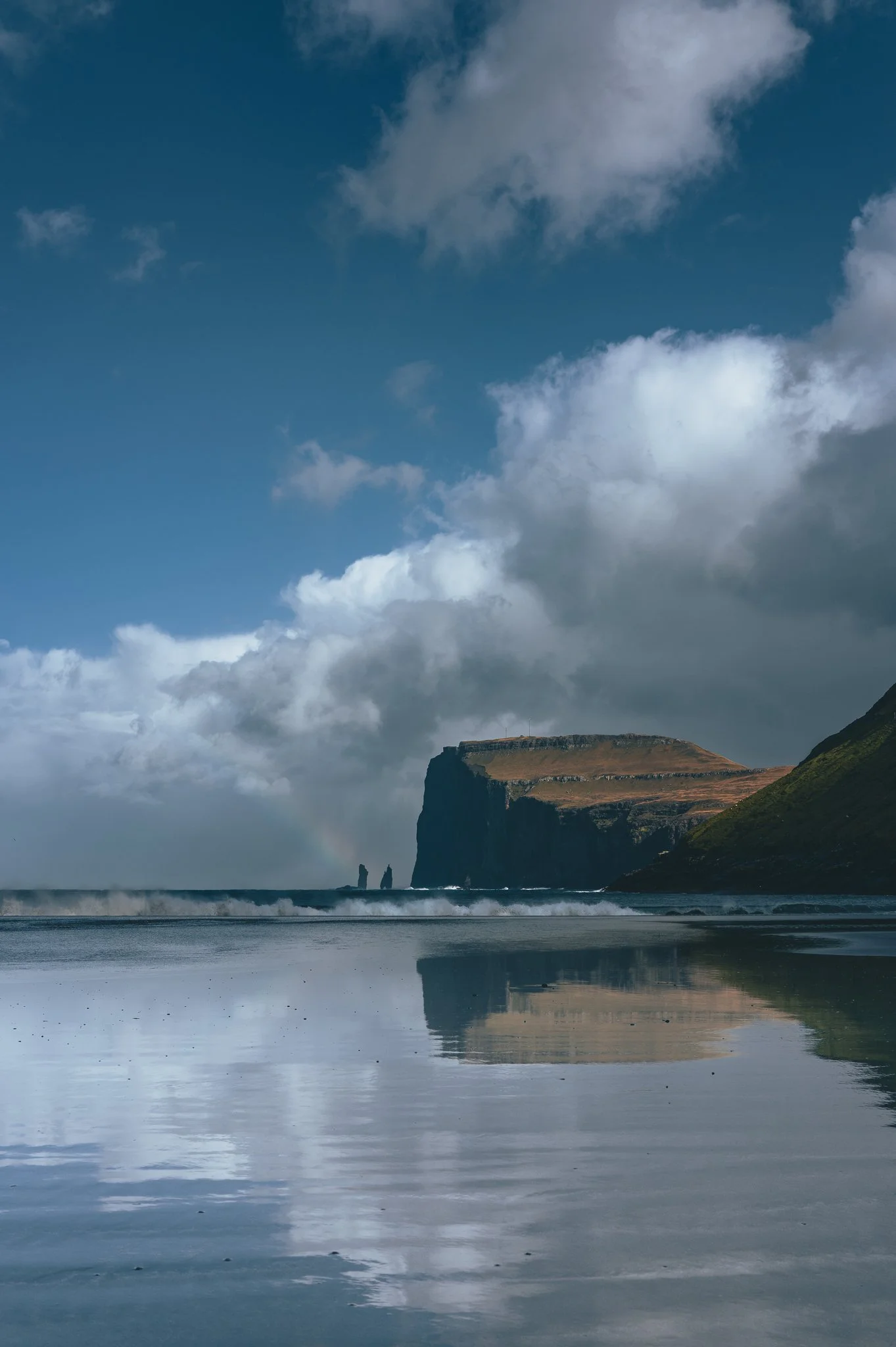 A coastal landscape with a cliff in the background under a cloudy sky and the reflection on the water.