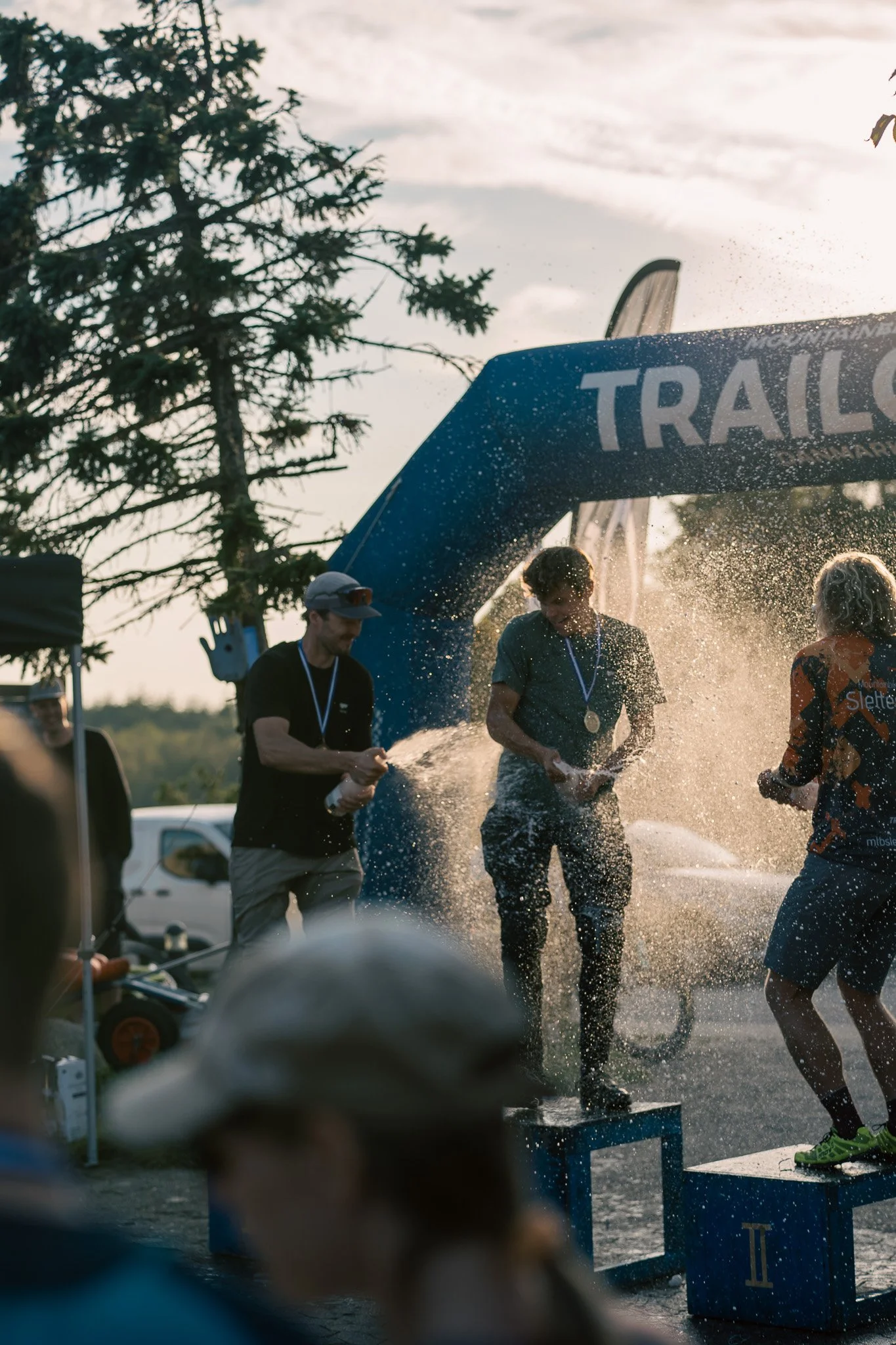 People celebrating on podium with medals, spraying each other with champagne at an outdoor event, with a large inflatable arch marked 'TRAIL' in the background, and a tree and vehicles visible in the distance during sunset.