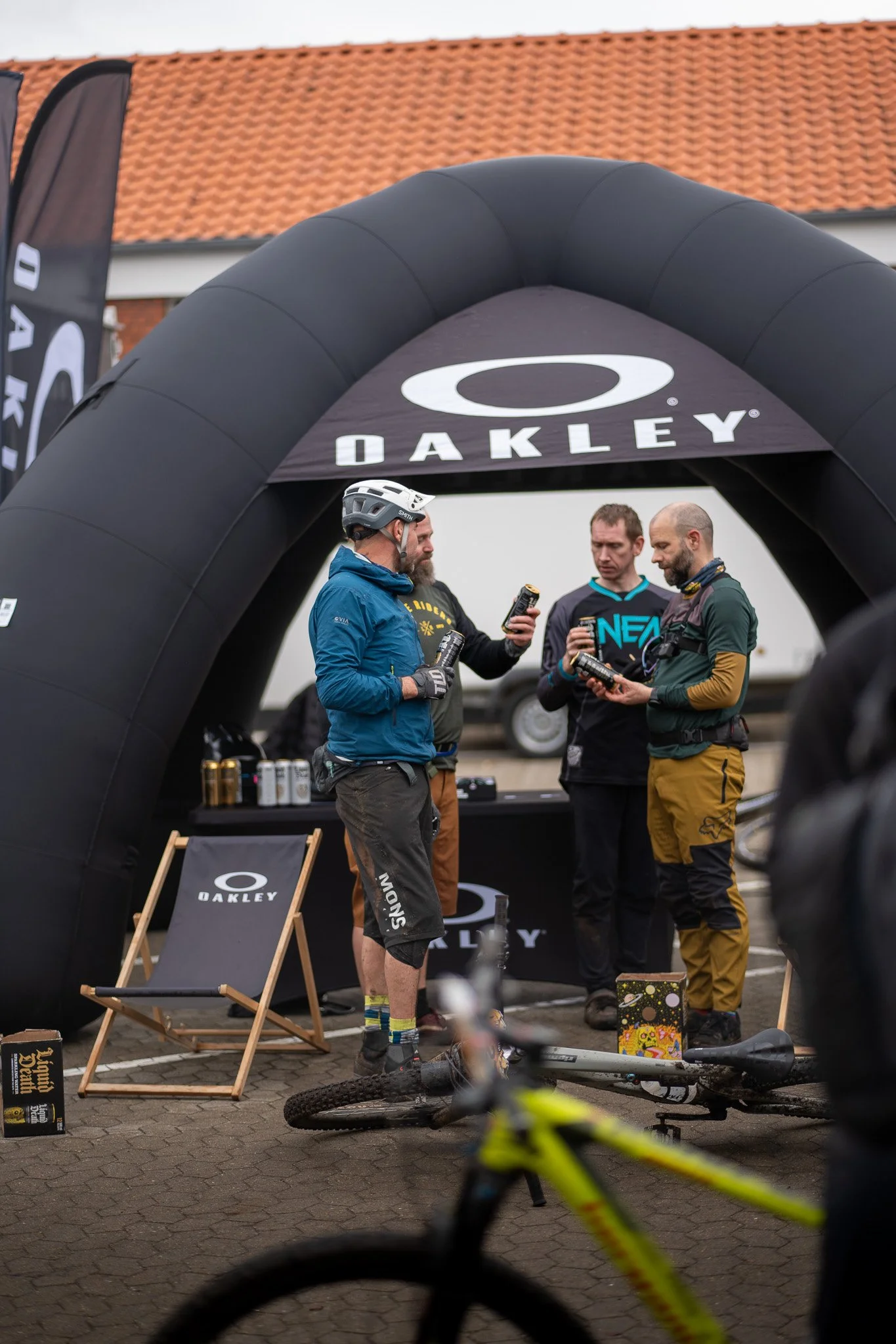 Four men at an Oakley event, with one wearing biking gear and helmet, chatting and holding cans under a black inflatable arch with Oakley branding. A mountain bike and promotional items are visible.