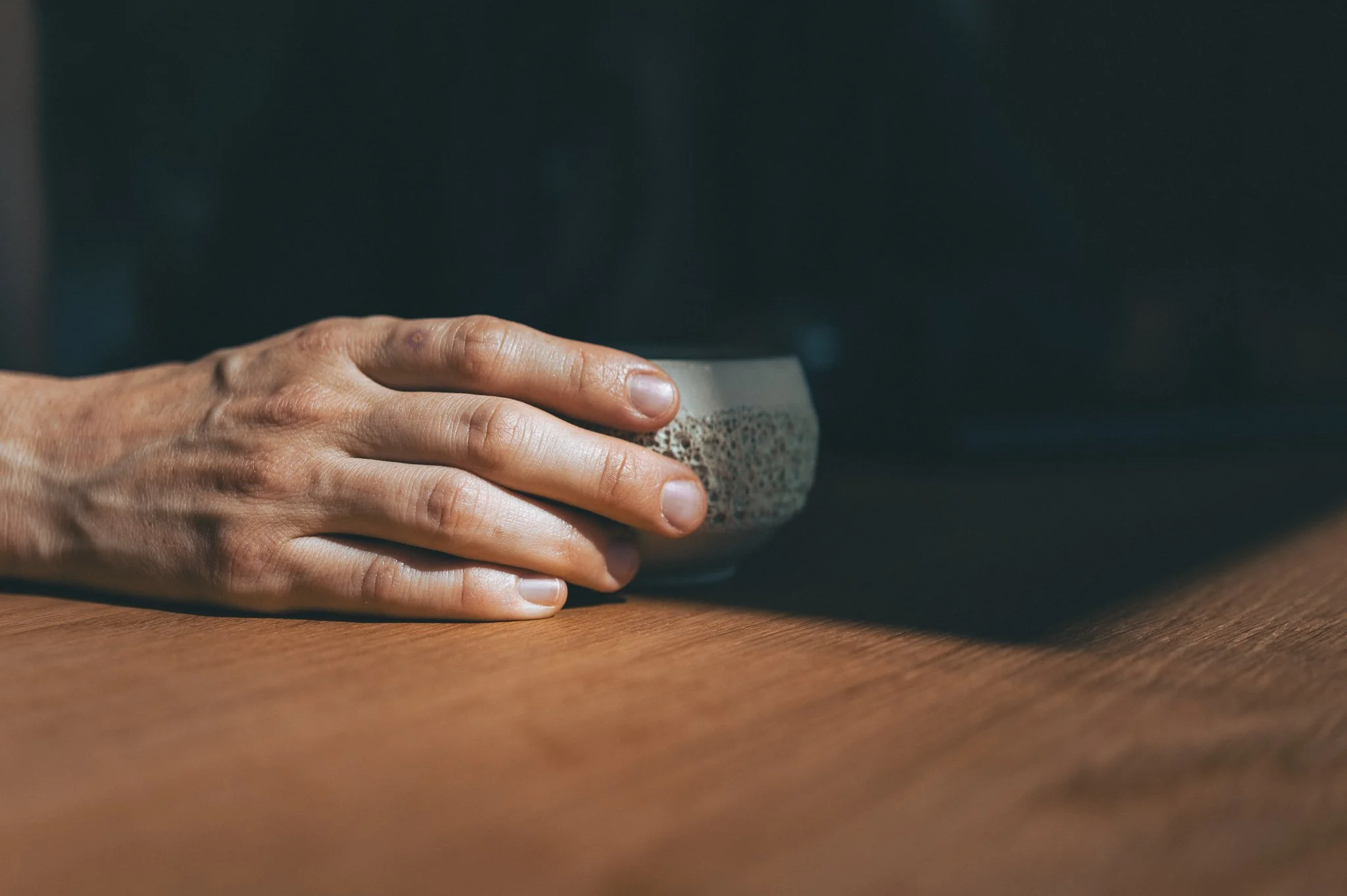 A person's hand resting on a wooden table, holding a ceramic mug.