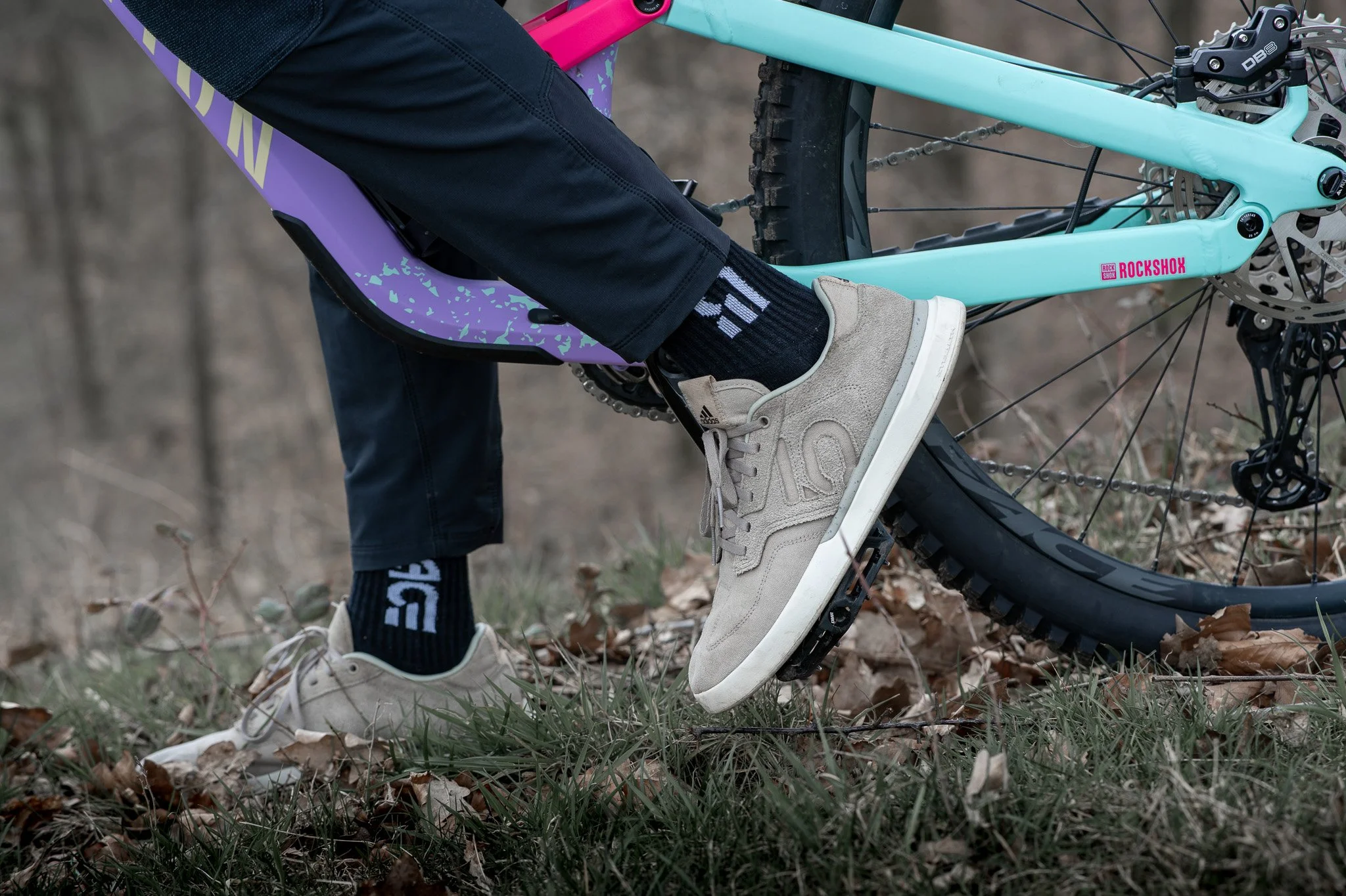 Close-up of a person wearing beige sneakers and black socks with white text, standing next to a light blue mountain bike on a grassy and leaf-covered ground in a wooded area.