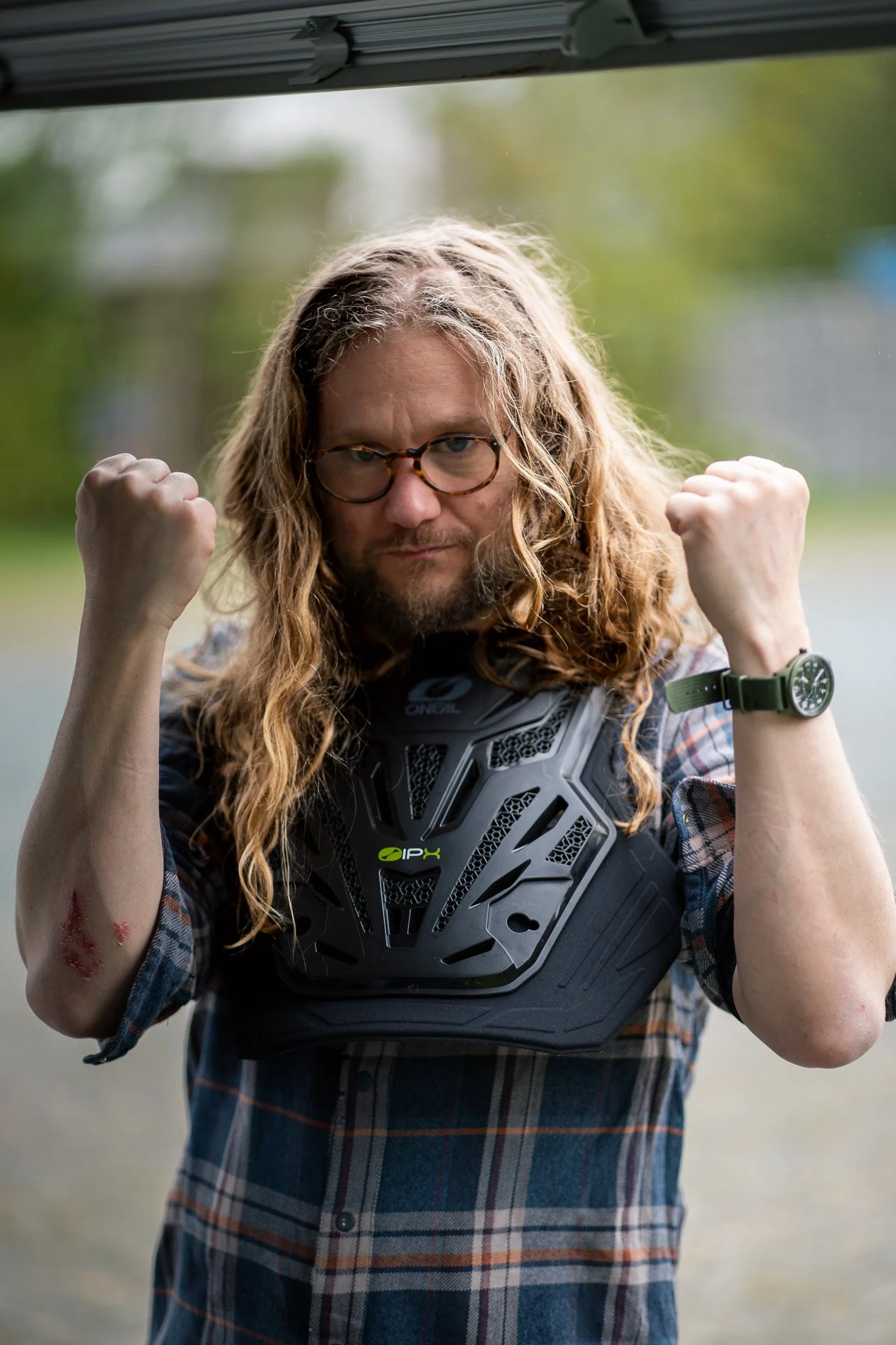 Man with long hair and glasses flexing his arms, wearing a protective chest armor, standing outdoors.