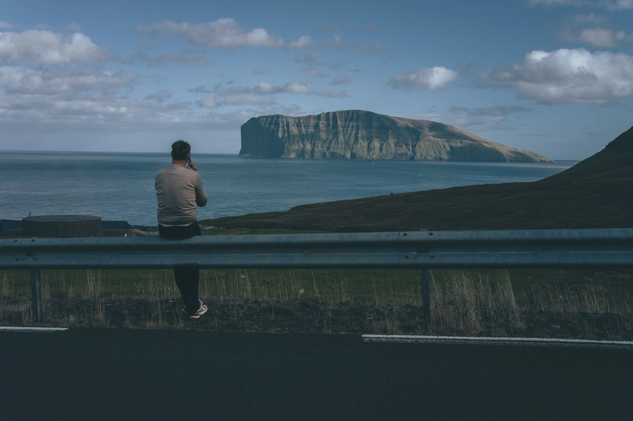 A man standing by a highway guardrail taking a photo of a large cliff or island in the ocean with a partly cloudy sky.