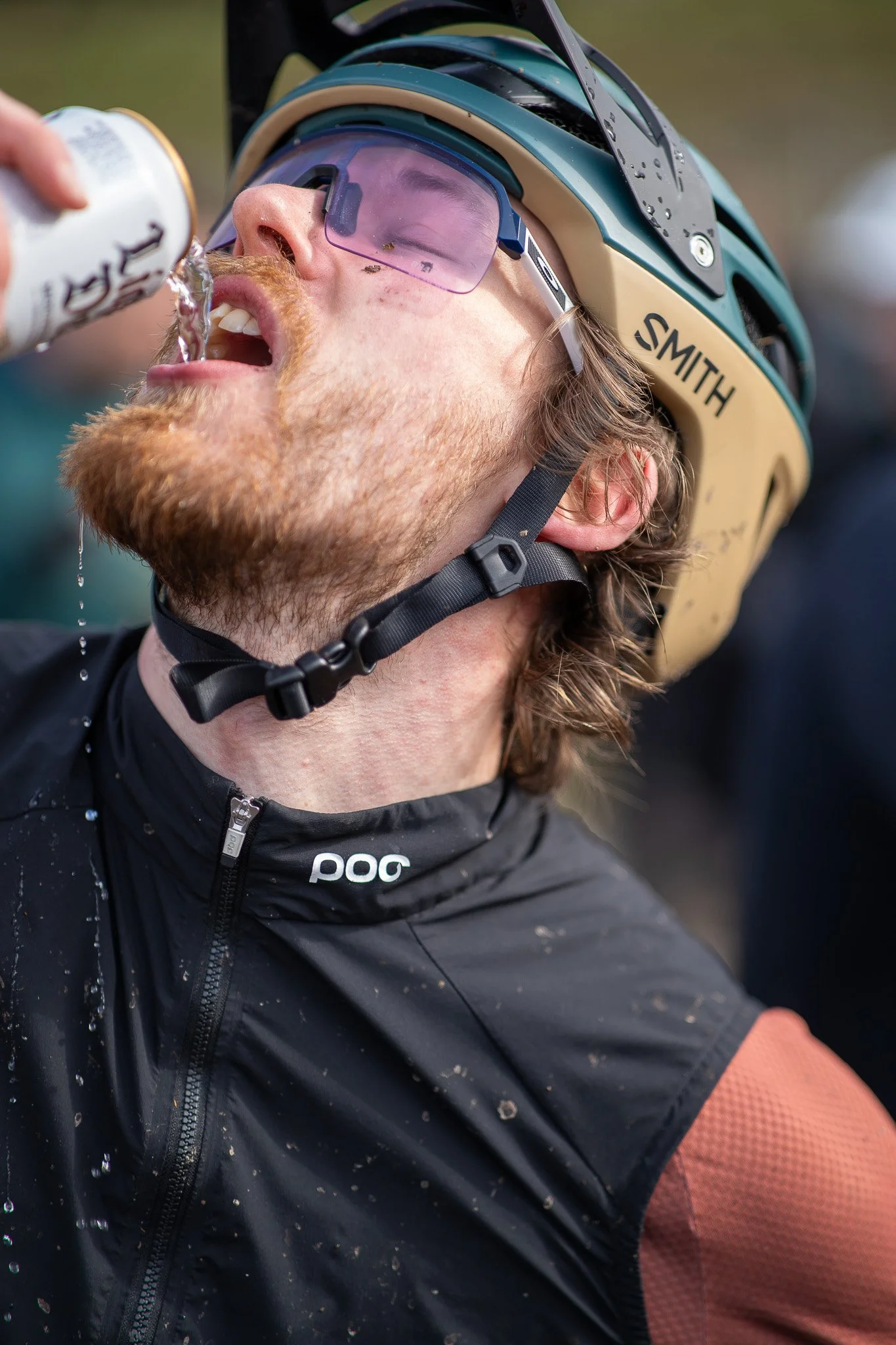 A man wearing a cycling helmet and sunglasses drinking water from a bottle during a rest.
