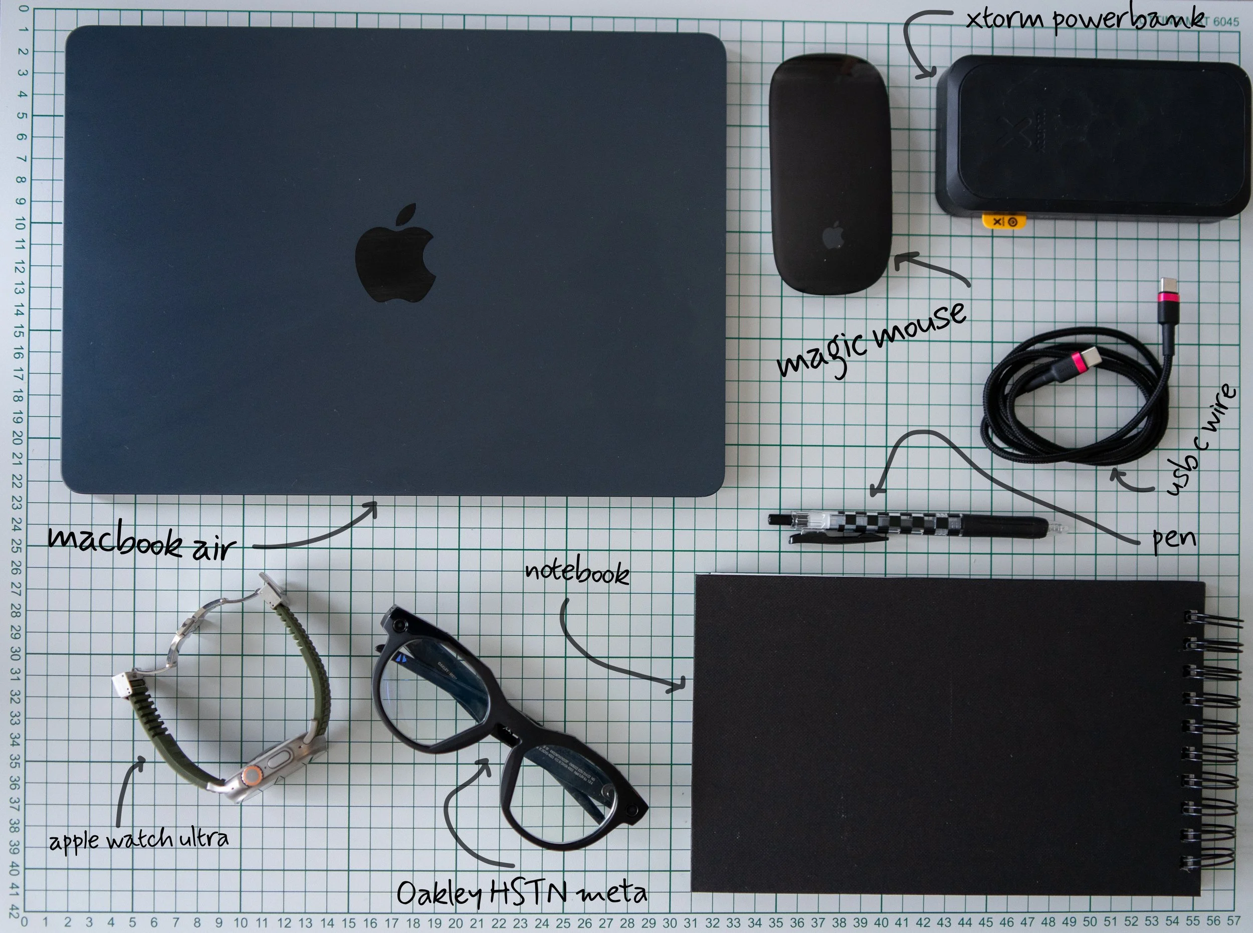 A flat lay of electronic and office items on a grid paper background. Includes a closed MacBook Air, the note books, a black Magic Mouse, a black Xtorm Power Bank, a black external hard drive, a USB-C cable, a checkered pen, and an Apple Watch Ultra with a white band.