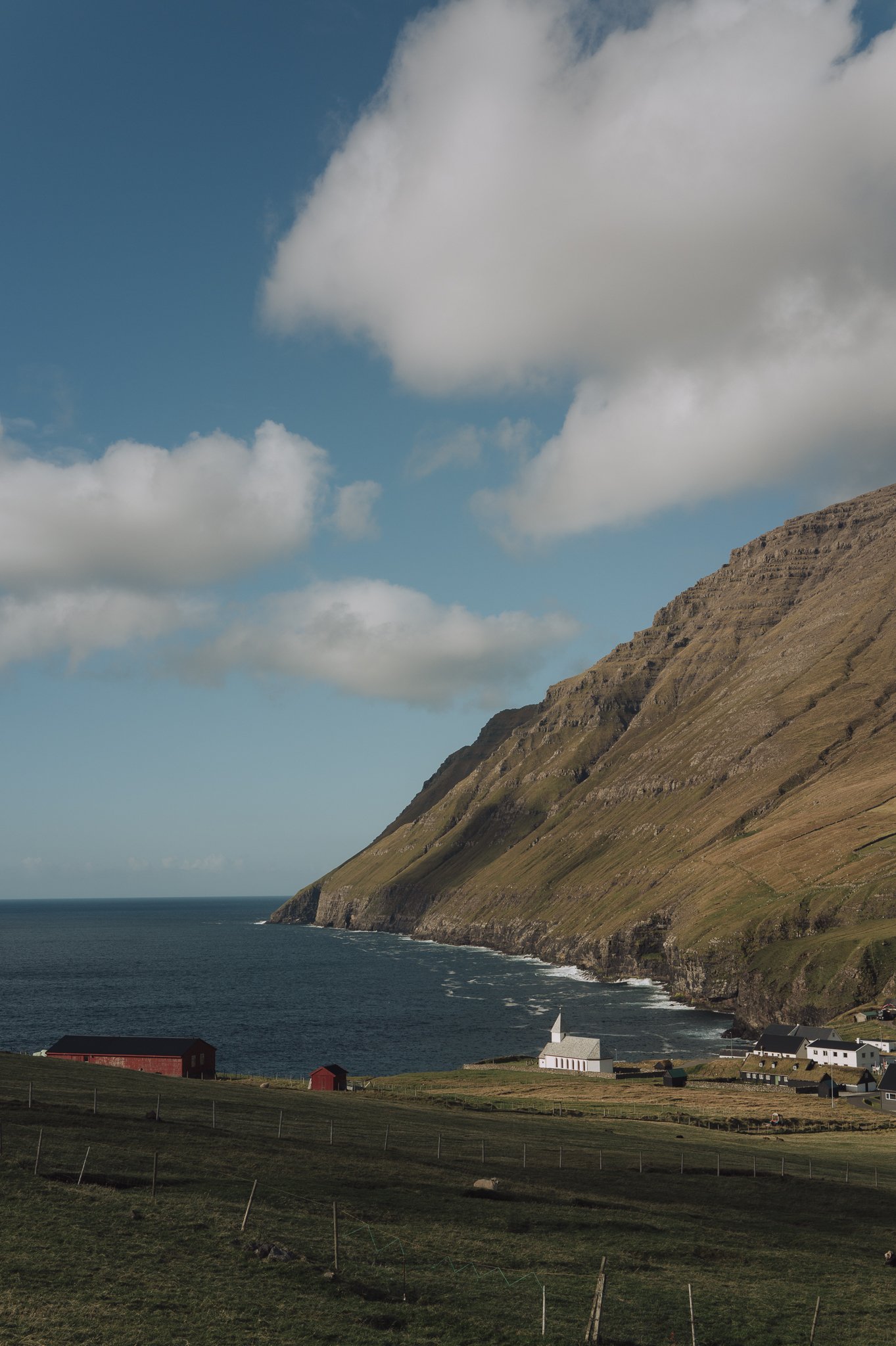 Coastal scene with a rugged hillside, a few scattered houses near the shoreline, and a large cloud in the blue sky.