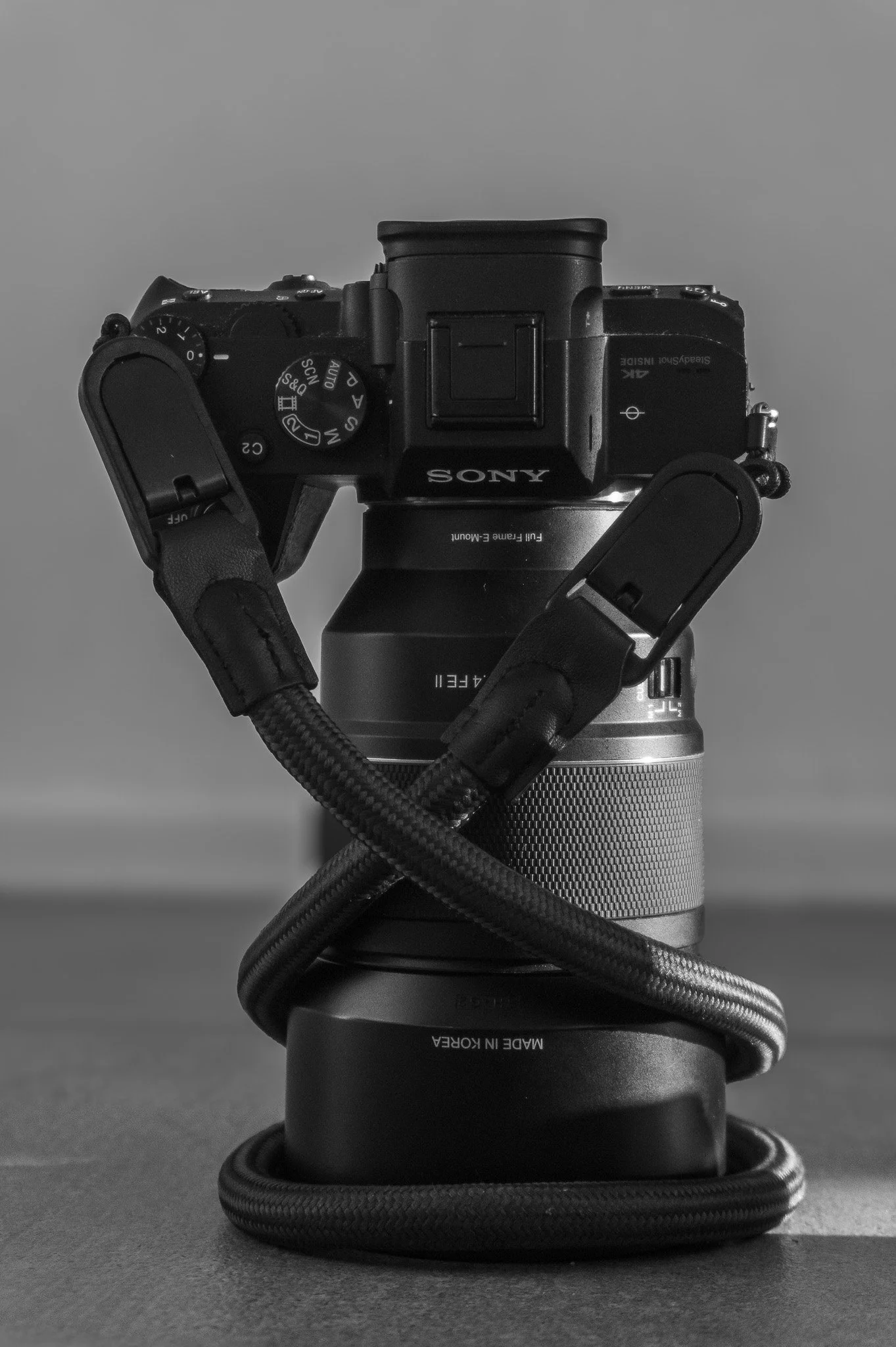 A black and white photograph of a Sony digital camera mounted on top of a camera lens, with a small camera strap looped around the lens. The camera is positioned against a blurred, plain background.
