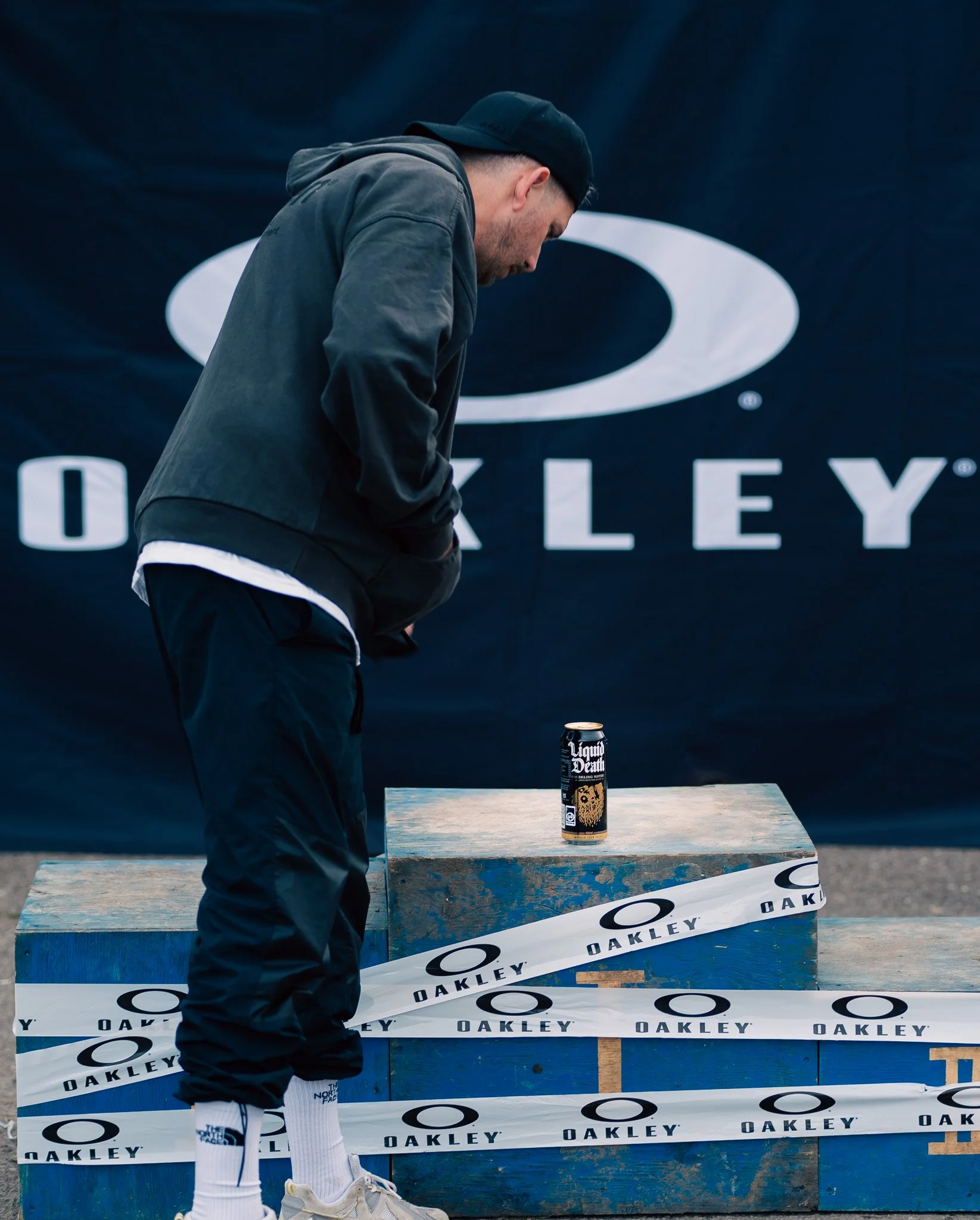 A man wearing a black hoodie, black pants, white sneakers, and a black cap looking down at a can of Liquid Death sparkling water on a blue wooden platform, with an outdoor Oakley backdrop.