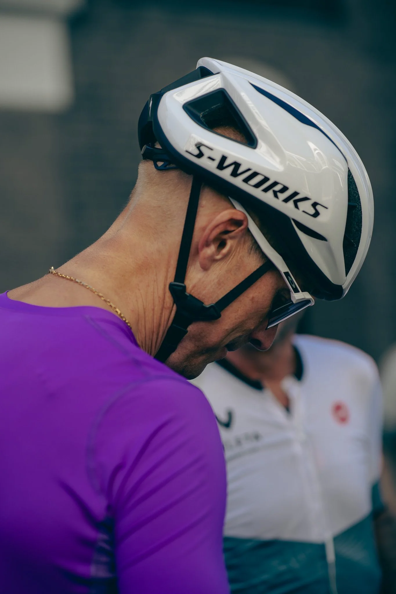 Close-up of a cyclist wearing a white S-Works helmet, sunglasses, and a purple cycling jersey, captured from the side with another person in the background.