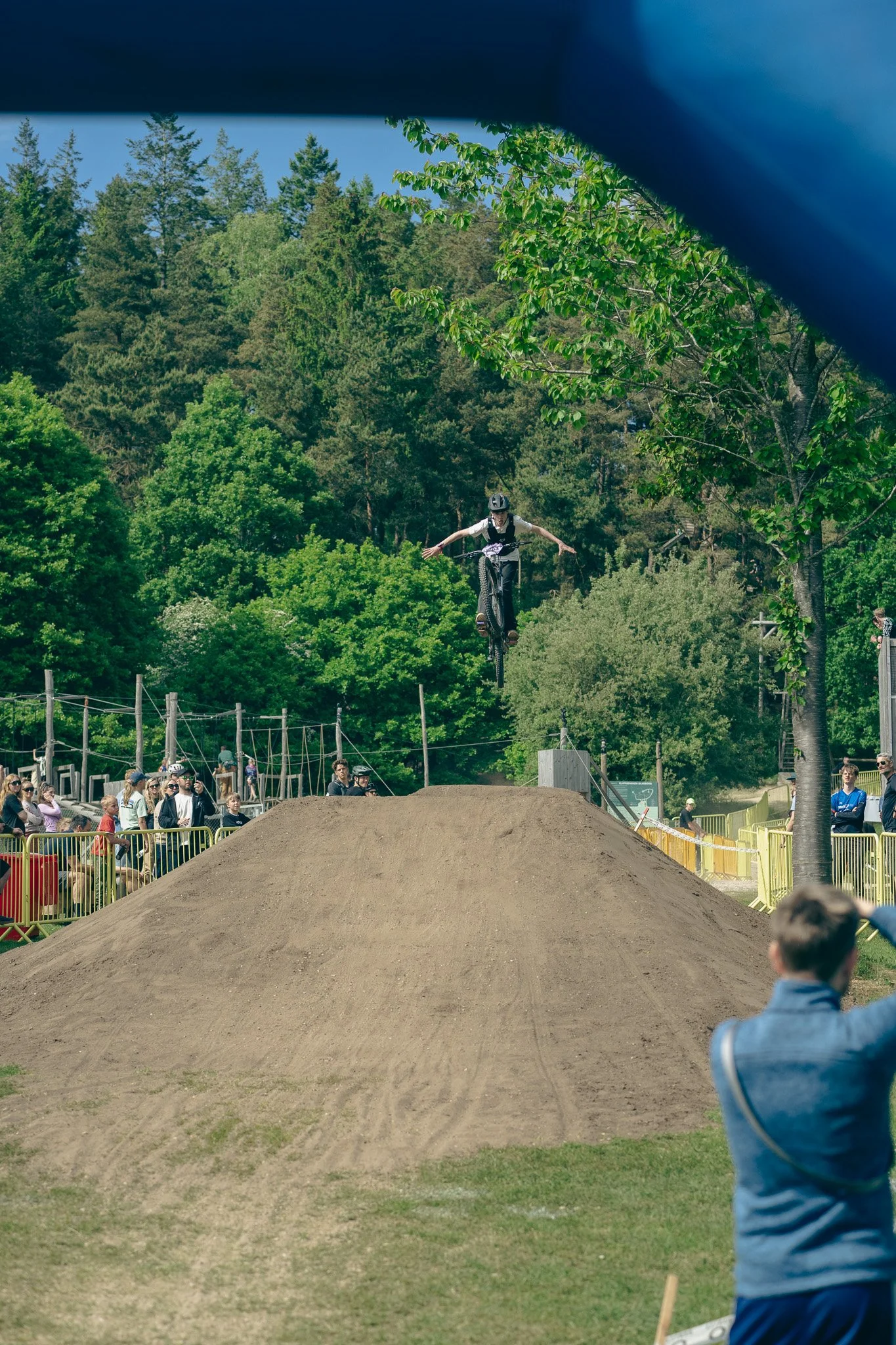 A person performing a bicycle jump off a dirt ramp during a bike race, with spectators watching along the sides.