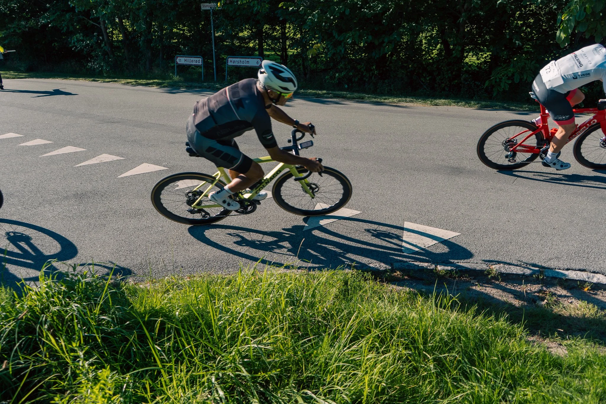 Two cyclists riding on a paved road with greenery on the side, with shadows visible on the road and road signs in the background.