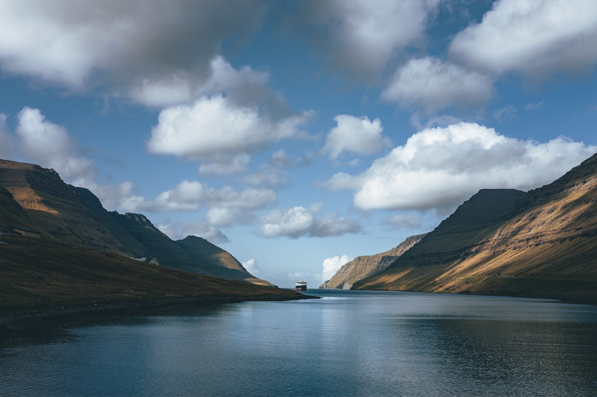 A narrow waterway flanked by green and rocky hills under a partly cloudy sky, with a large ship sailing in the distance.