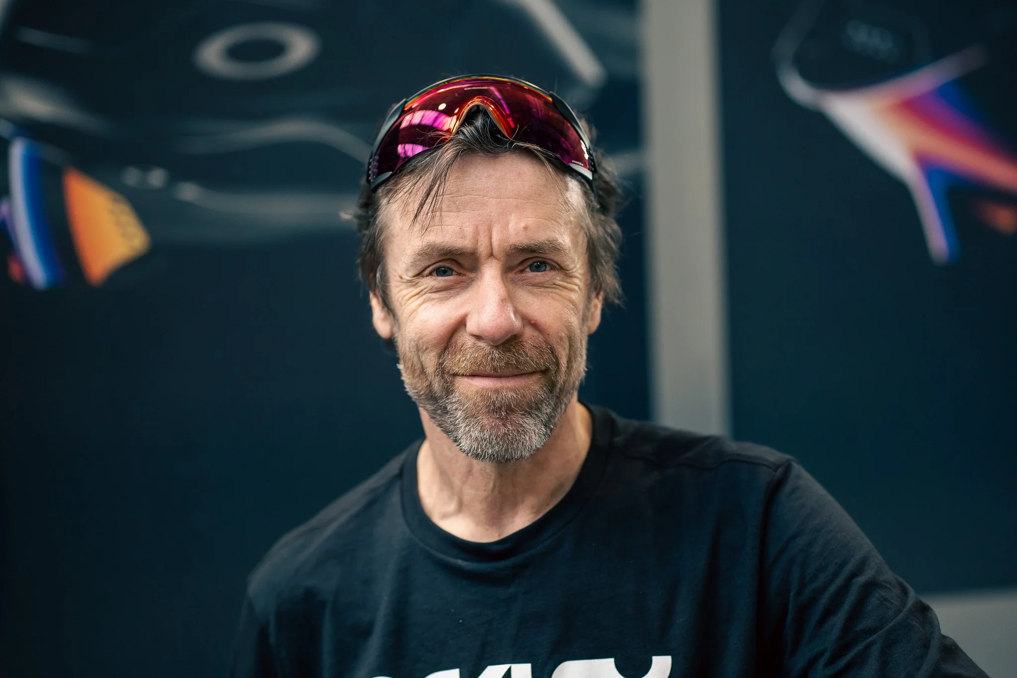 A middle-aged man with a beard and short hair, wearing sunglasses on his head and a black t-shirt, smiling at the camera with a dark blue background and abstract shapes.