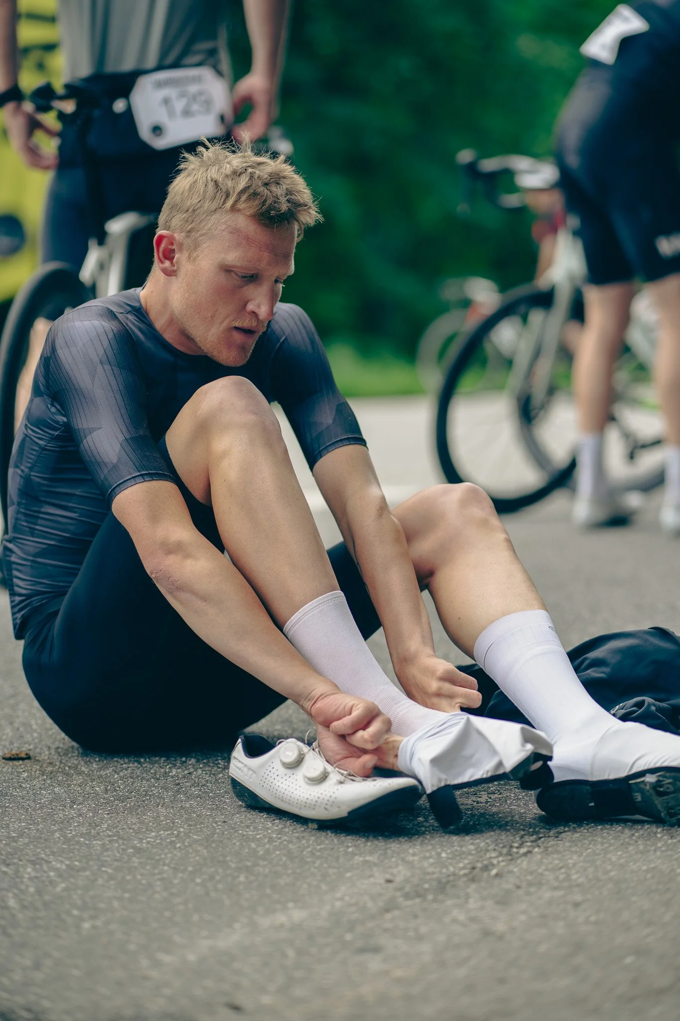 A male cyclist sitting on the ground adjusting his cycling shoes after a race, with two other cyclists and bicycles in the background.