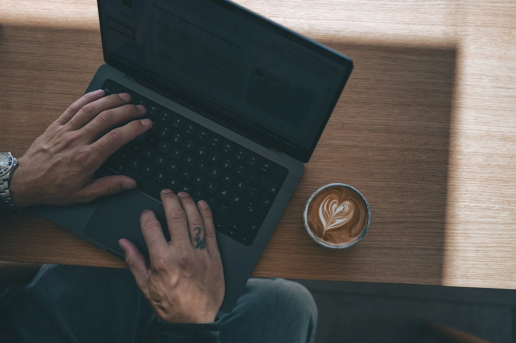 A person with tattoos on their hands is using a laptop on a wooden table. Next to the laptop, there is a cup of coffee with a heart-shaped latte art on top.