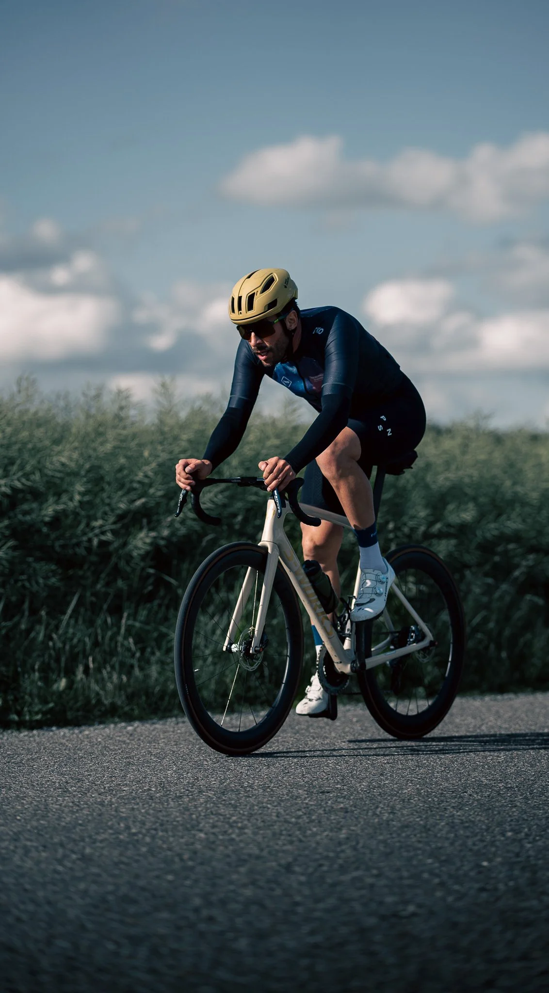 A cyclist riding a road bike on a paved path through a rural area with green bushes and a cloudy sky.