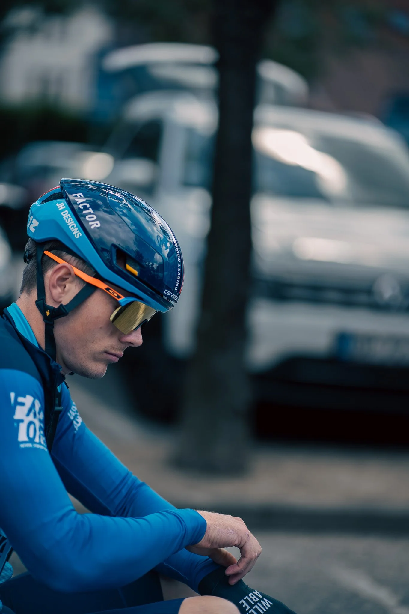 A male cyclist wearing a blue helmet, sunglasses with golden lenses, and a blue cycling jersey sitting on a bike with a serious expression, with parked vehicles in the background.