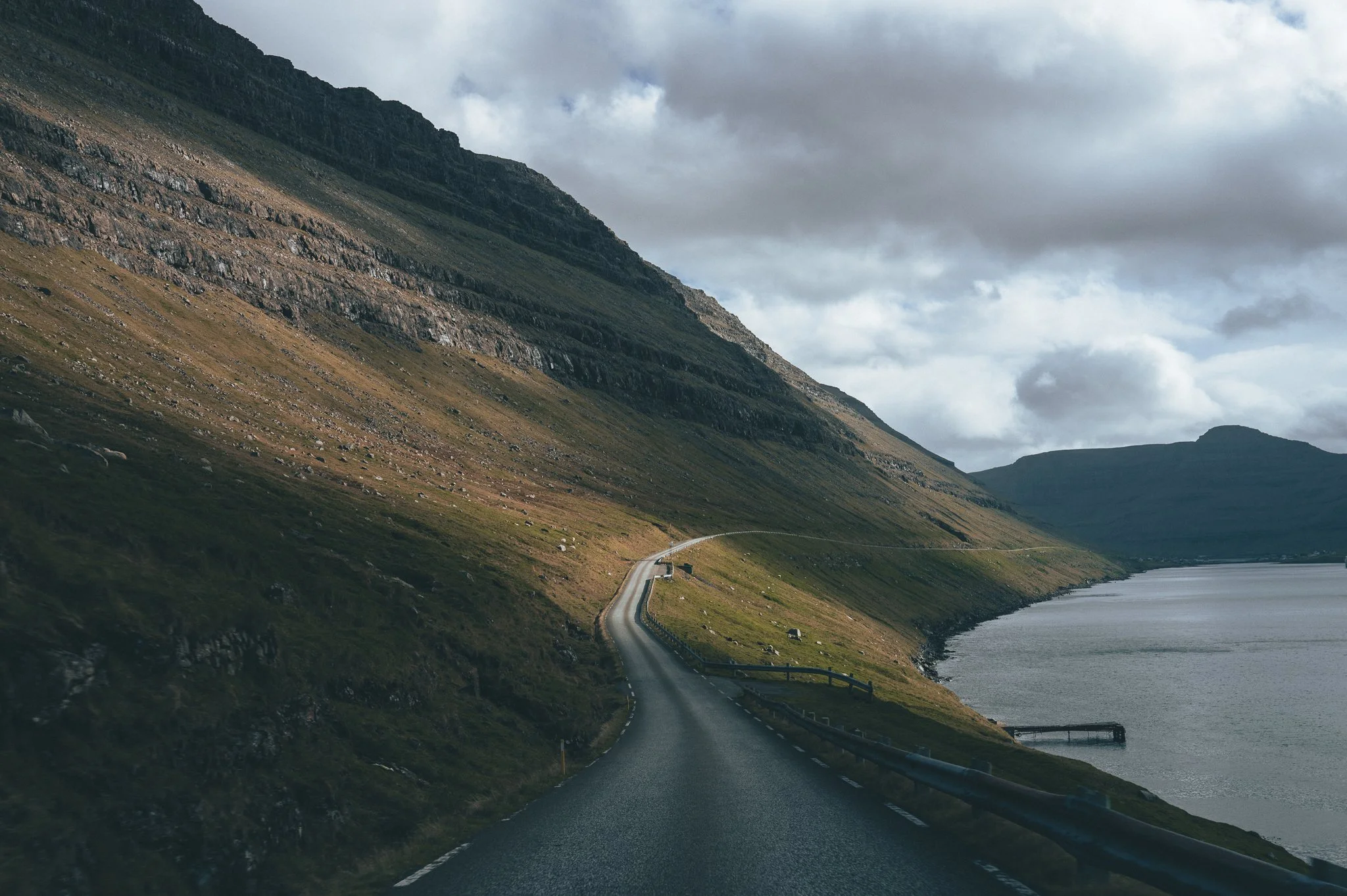A winding coastal road runs along the edge of a body of water, with steep, grassy, and rocky mountains on one side and cloudy skies overhead.