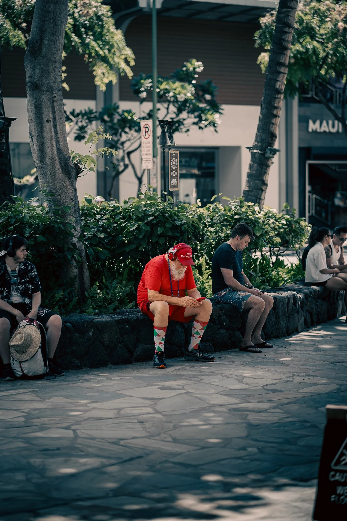 People sitting on a stone bench in an outdoor shopping area, including an older man dressed as Santa Claus with a red hat, red shorts, sporting Argyle socks, and using a smartphone.